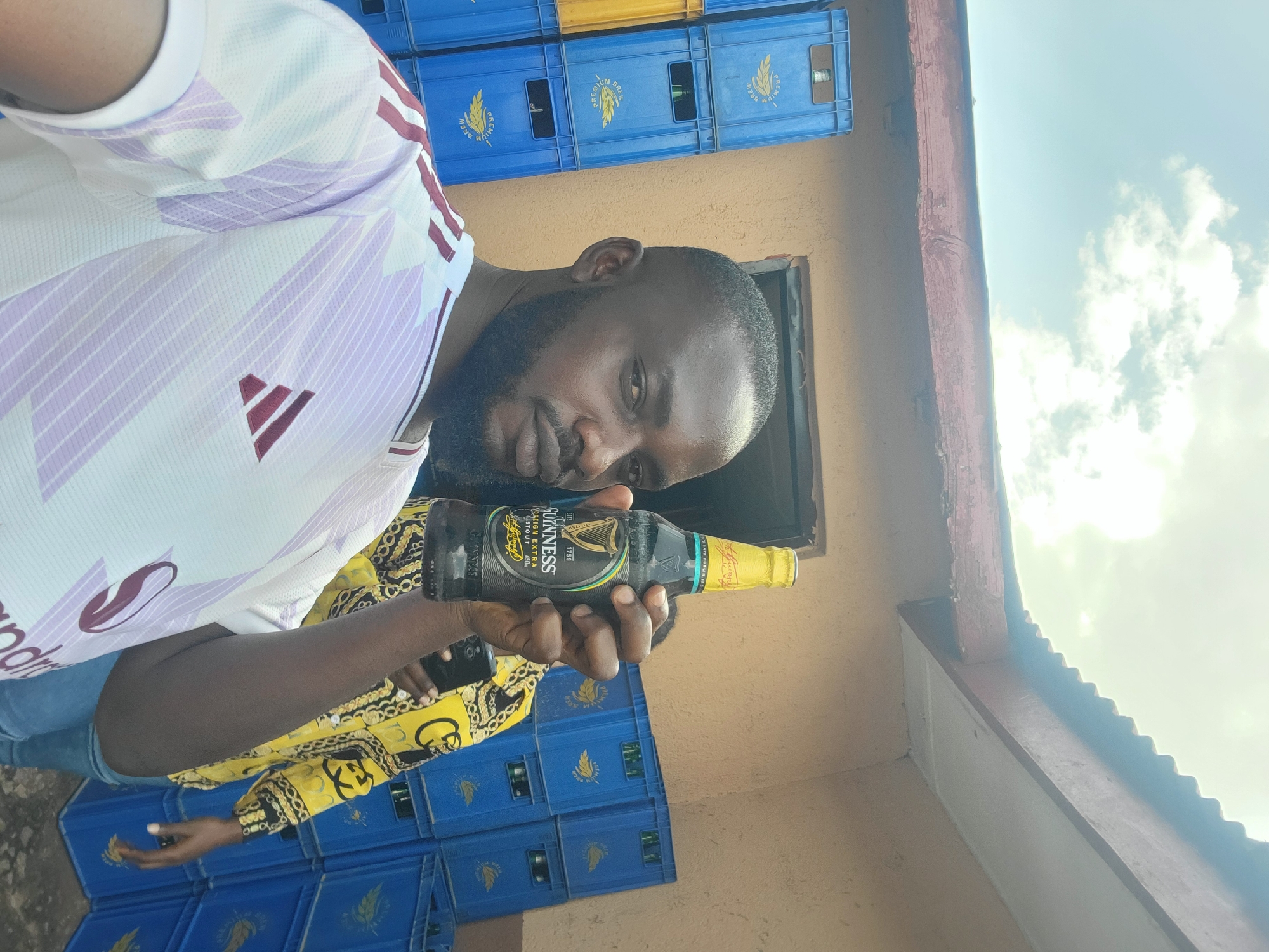 A man is holding a bottle of Guinness Foreign Extra Stout in front of a wall of blue crates, some of which are filled with bottles. The man is wearing a white t-shirt with a red Adidas logo and purple stripes, and a yellow shirt with a black and gold pattern. He has a beard and is looking towards the camera. Behind him, stacks of blue and yellow crates are visible. The sky is cloudy. The image appears to have been taken outdoors in Jos, Nigeria. The lighting suggests it is daytime. The man's expression is neutral. Text on the Guinness bottle is visible and reads "GUINNESS FOREIGN EXTRA STOUT". Other text on the crates reads "PREMIUM BREW".