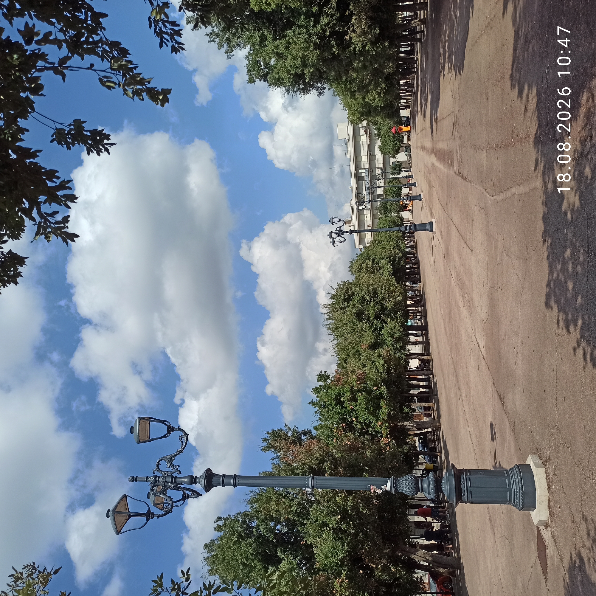 A wide shot captures a paved public square in Bitonto, Italy, under a bright blue sky with scattered white clouds. A prominent, ornate dark gray lamppost stands on the left side of the foreground, its detailed base resting on a light-colored hexagonal platform. In the background, a row of mature trees lines the perimeter of the square, with a multi-story building visible behind them. The pavement is marked with cracks and shows signs of wear. A few people are present in the mid-ground, appearing to be walking or standing near benches. Further in the distance, playground equipment, including a red slide, is visible. The bottom right corner of the image displays a timestamp: "18.08.2026 10:47".