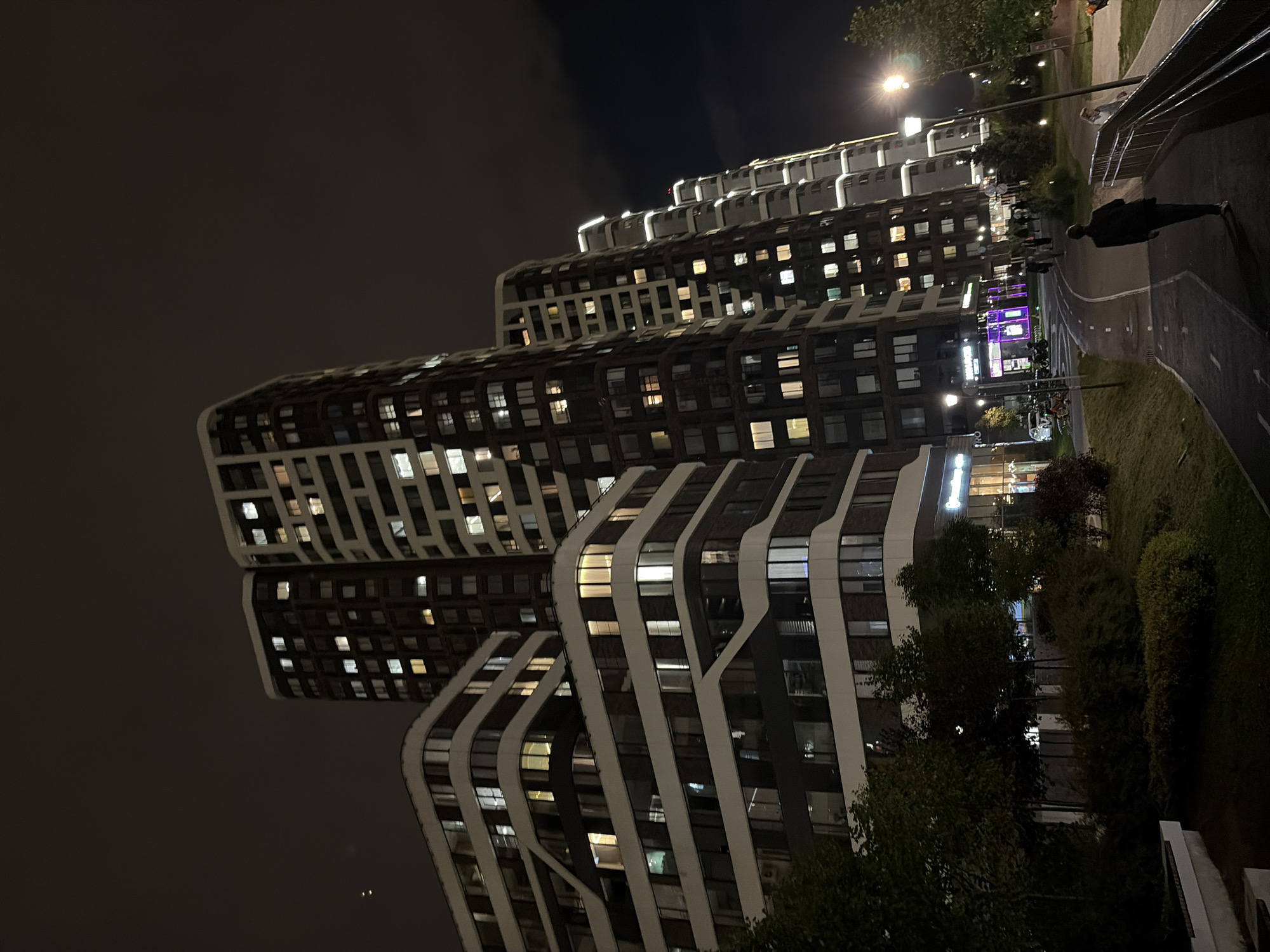 This image depicts a modern residential complex at night in Moscow, Russia. The architecture features several tall, contemporary buildings with numerous illuminated windows, indicating residential occupancy. The buildings have distinct facade designs; one appears to have a white, wavy pattern against a dark background, while others are more rectilinear with a mix of dark and lighter panels. The scene includes a pedestrian walkway with a visible lane marking and a subtle gradient, bordered by green spaces and trees. Streetlights illuminate the path. In the foreground, a lone figure walks away from the camera on the pathway. The sky is dark and cloudy, characteristic of an evening or night in an urban environment. The overall impression is one of a well-lit, developed urban residential area.