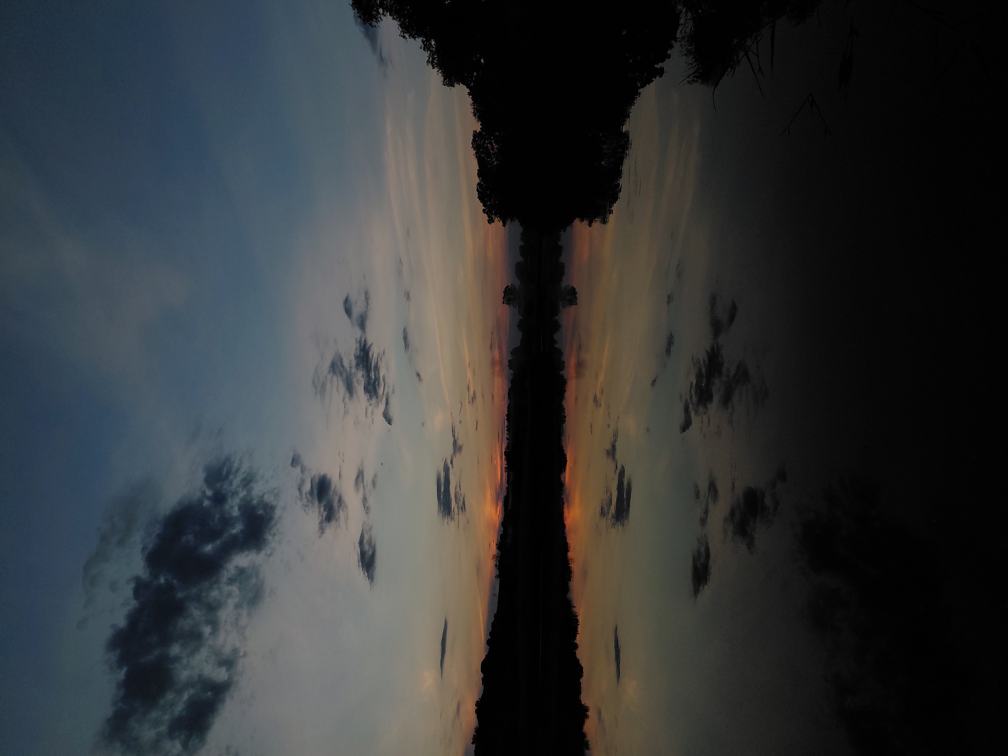 This image, captured in Ustyvytsia, Ukraine, depicts a tranquil twilight scene over a calm body of water, likely a river or lake. The sky transitions from a deep, muted blue at the top to lighter shades of grey-blue, fading into warm orange and pink hues along the horizon, suggesting either sunrise or sunset. Scattered dark grey clouds are present across the upper sky and near the horizon. A dark, silhouetted landmass, consistent with a distant bank or hills adorned with trees and vegetation, defines the horizon line. The placid water surface perfectly reflects the sky's colors and cloud formations, mirroring the light and dark elements with distinct clarity. No human subjects, man-made structures, or explicit actions are discernible within the frame; the scene is entirely natural, characterized by its low light and reflective qualities.