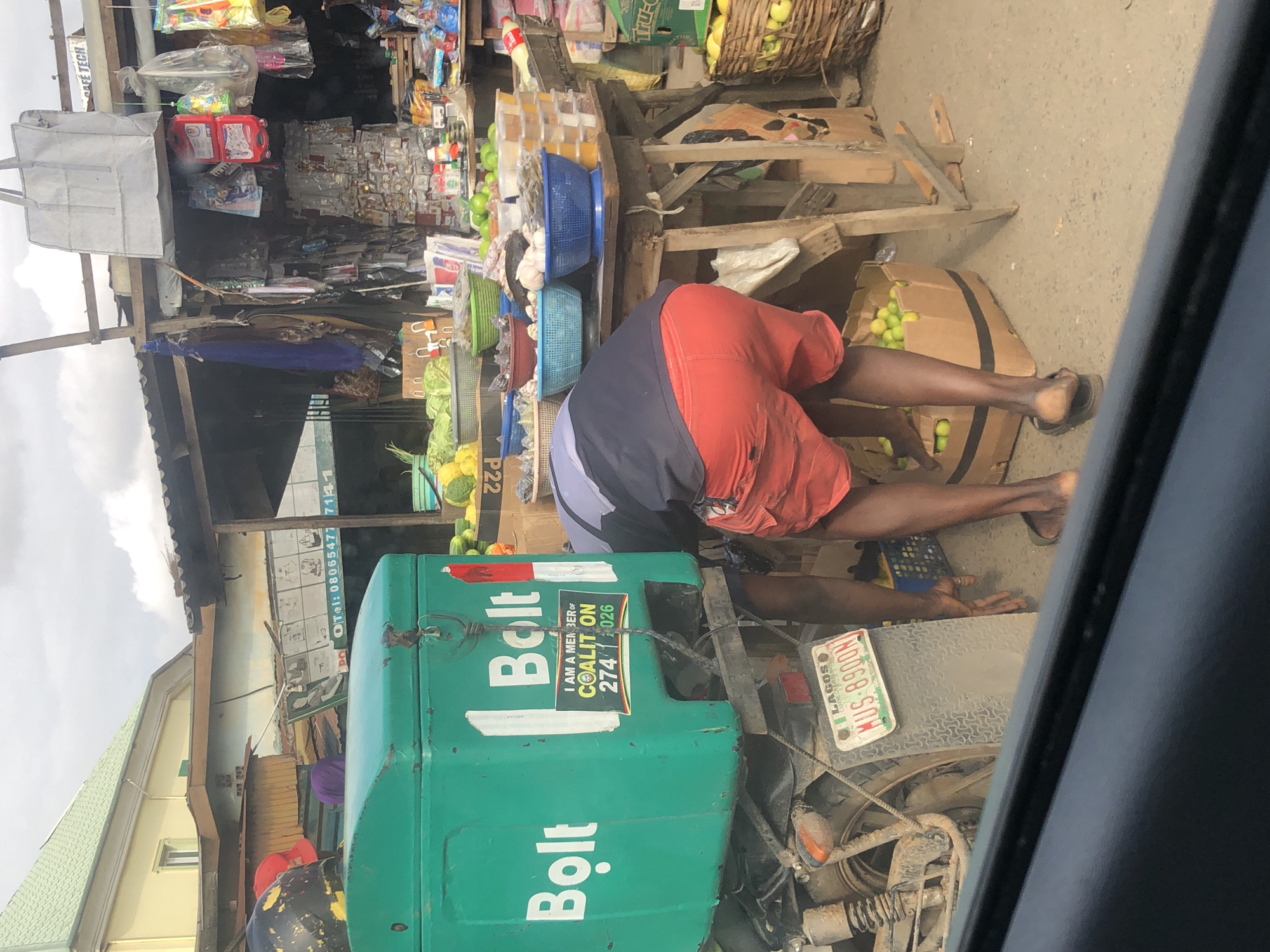 The image depicts a street scene in Lagos, Nigeria, with a focus on a vendor at a market stall. A person wearing red shorts and a grey and black shirt is bent over, attending to a stall laden with produce and various goods. The stall itself is constructed from wood and corrugated metal, typical of informal market structures. In the foreground, a green delivery box with the "Bolt" logo is mounted on a motorcycle. A license plate reading "LAGOS KUS-8900N" is visible on the motorcycle. Various fruits, vegetables, and packaged items are displayed on the stall. The ground is paved with concrete, and the background shows more stalls and buildings, suggesting a busy marketplace environment. The lighting indicates it is daytime.