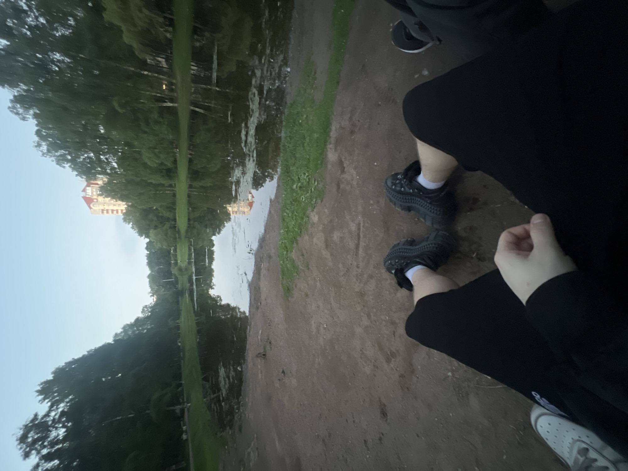 The image is a low-angle, first-person perspective photograph depicting an outdoor scene from the viewpoint of a seated individual. In the immediate foreground, the lower legs and feet of two individuals are visible, resting on a brown dirt ground. The person taking the photograph wears black shorts or pants; their left hand rests on their left thigh, and their right foot is clad in a black, chunky-soled sneaker with a white sock. A light-colored sneaker is partially visible on their left foot. To the right, the legs and black sneakers of a second person are partially visible.
Beyond the dirt foreground, a narrow strip of green grass borders a calm body of water, likely a pond or small lake. The water reflects the pale sky and surrounding trees, and scattered patches of aquatic vegetation are visible on its surface.
The background features a dense line of trees of various species, including some with light-colored bark, stretching along the far bank of the water. Above the tree line, towards the upper right, a multi-story building with a distinctive reddish-orange roof is partially visible, indicating an urban or suburban environment. The sky is a clear, light blue. The overall setting appears to be a park or green space located within or on the outskirts of the city of Unknown, Unknown, captured under soft, diffuse lighting conditions, possibly at dusk or dawn.