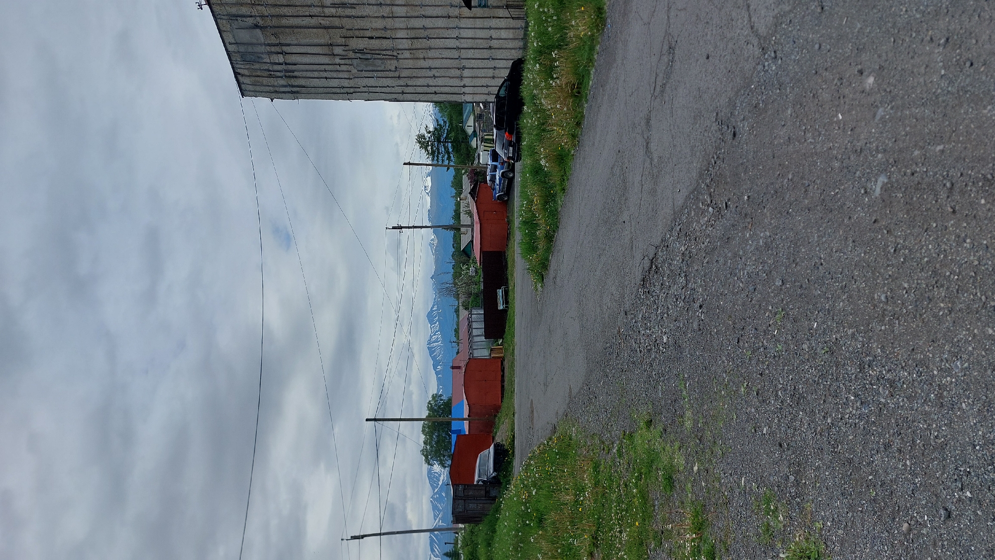 The image depicts a view of a village with snow-capped mountains in the background. The foreground shows a gravel road leading to a cluster of small, brightly colored buildings, likely garages or sheds, some with red walls and blue roofs. Several parked vehicles, including a dark-colored car and a blue SUV, are visible near the buildings. To the right, a large, textured concrete building dominates the scene, with visible vertical supports and a rough, weathered facade. The sky is overcast with grey clouds. The overall impression is one of a rural or semi-urban environment in a mountainous region, possibly during a cool or cloudy day. The location is identified as Зелёный, Russia.