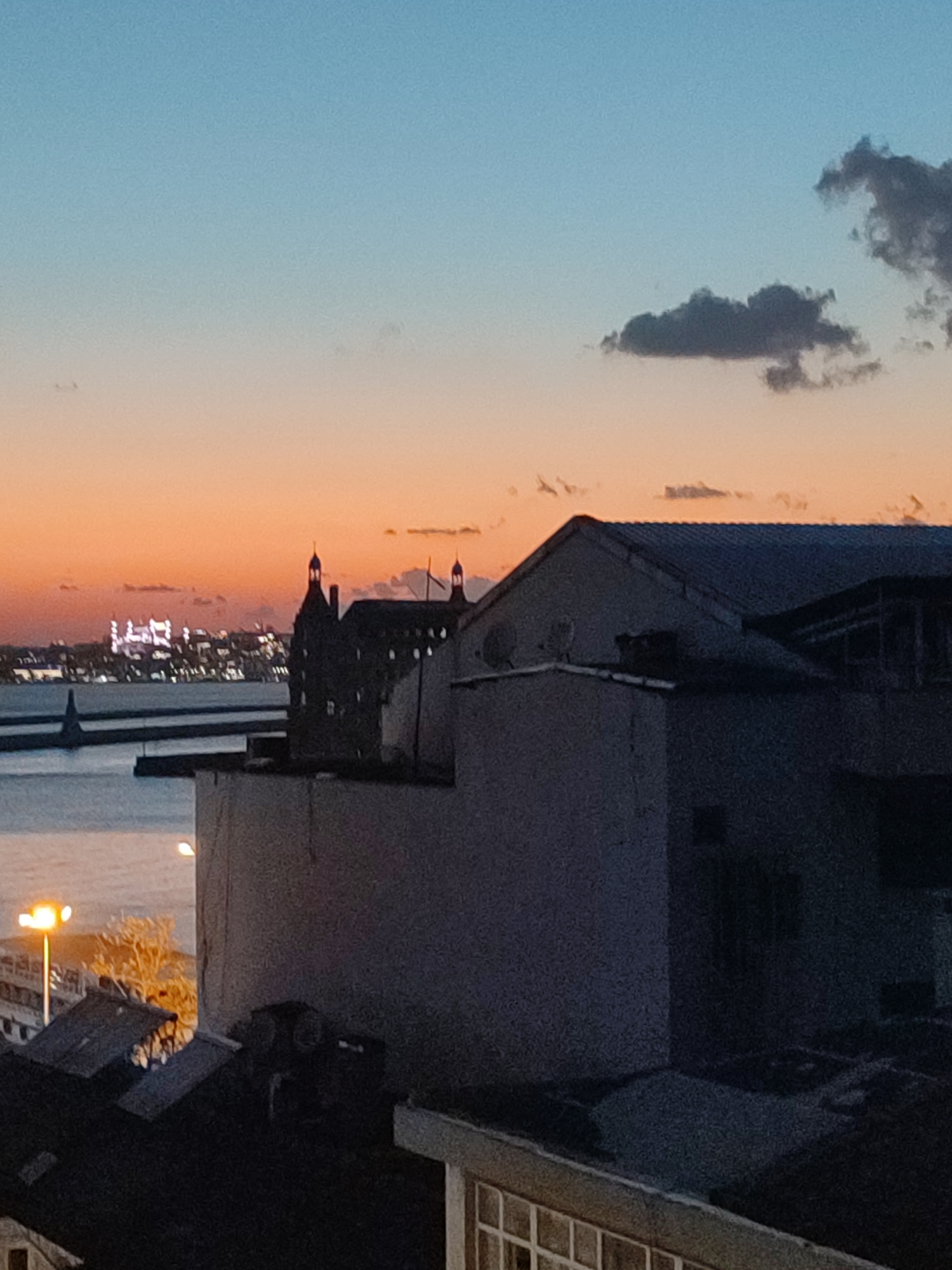 The image captures a dusk scene overlooking the Bosphorus Strait, likely from Kadıköy, Turkey, with the distant skyline of Istanbul. In the foreground, the dark silhouettes of rooftops and buildings dominate, obscuring most details. A large, partially visible structure with a tiled roof and what appears to be a balcony or an enclosed area is prominent in the right half of the frame. To the left of this structure, a lighter-colored wall recedes into the background. Further left and lower, a streetlamp with a bright orange light illuminates a barren tree with bare branches. A body of water, presumably the Bosphorus, stretches across the mid-ground, bordered by a dark, low-lying structure resembling a pier or breakwater. On the opposite side of the water, the illuminated skyline of Istanbul is visible, with distinct buildings and structures highlighted by lights, including what appears to be a large, white, multi-story building. The sky transitions from a deep blue at the top to a warm orange hue near the horizon, with a few dark clouds scattered across the upper part of the frame.