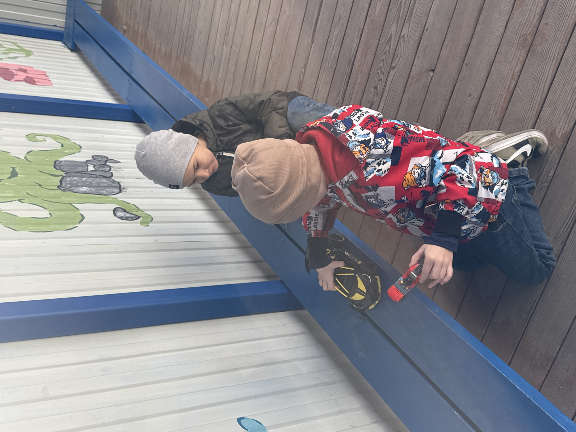 The image shows two young boys playing with toy cars on a blue beam, set against a backdrop of a wooden deck and a white corrugated wall. The location is Yekaterinburg, Russia.
One boy, wearing a grey beanie and a dark green jacket, is lying on the wooden surface, looking down at the blue beam. Beside him, another boy, dressed in a light brown beanie, a red jacket patterned with cartoon characters (resembling Paw Patrol), and blue jeans, actively manipulates two toy cars. He holds a black and yellow toy car in one hand and a smaller red toy car in the other, guiding them along the blue structure.
The background features a wooden deck and a white corrugated wall with playful green and pink painted designs, including what appears to be a large green octopus or squid and pink floral elements. The blue beams provide a track-like surface for their play. The lighting is soft and even, suggesting daytime conditions, possibly overcast or indoors. The boys appear focused and engrossed in their activity. Visible text on the red jacket repeatedly shows "MISHA LASS" or "MISHA CLASS".