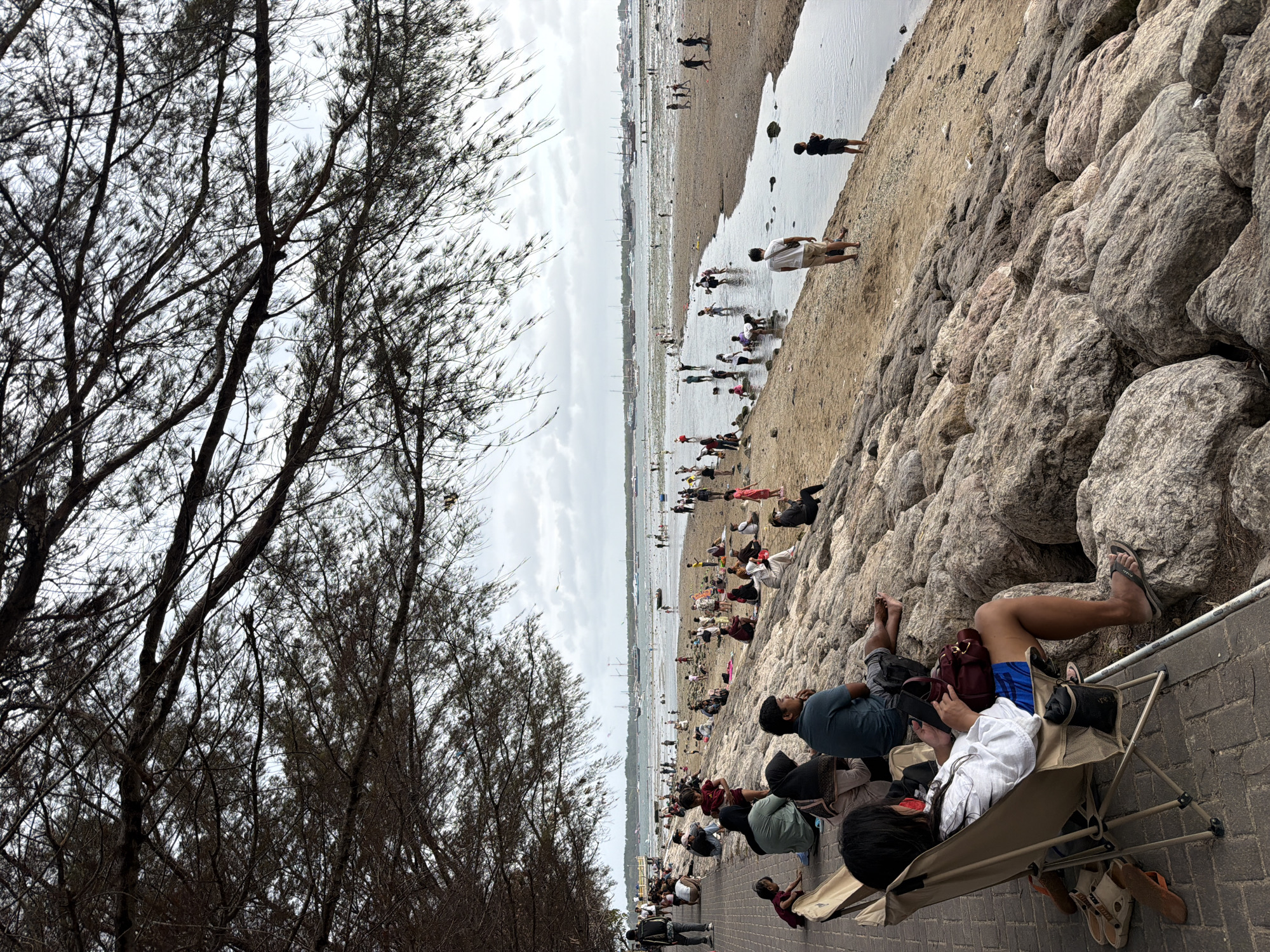 The image depicts a scenic view of Sanur Beach in Denpasar, Indonesia, on a cloudy day. A large group of people are spread across the sandy shore and the rocky embankment. In the foreground, several individuals are seated on chairs and rocks, observing the beachgoers and the ocean. Among them, a woman in a white shirt and blue shorts is engrossed in her phone, seated in a portable chair. Further down the embankment, more people are scattered, some sitting and others standing.
The beach itself is bustling with activity. People are wading in the shallow water, playing, and interacting with each other. A small river or stream cuts through the sand, leading to the sea. In the distance, several boats are anchored in the calm waters of the bay, with a cityscape visible on the horizon. Overhanging branches from a tree create a natural frame for the scene, casting shadows and adding depth to the composition. The overall atmosphere appears relaxed and leisurely, with many enjoying the coastal environment.