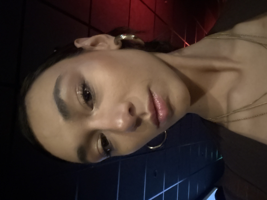 A person poses at an angle, their face prominently displayed. They have a gold earring and multiple delicate gold necklaces. The background appears to be a wall, with a darker portion below and a reddish section above. The lighting is dim and focused, highlighting their features.