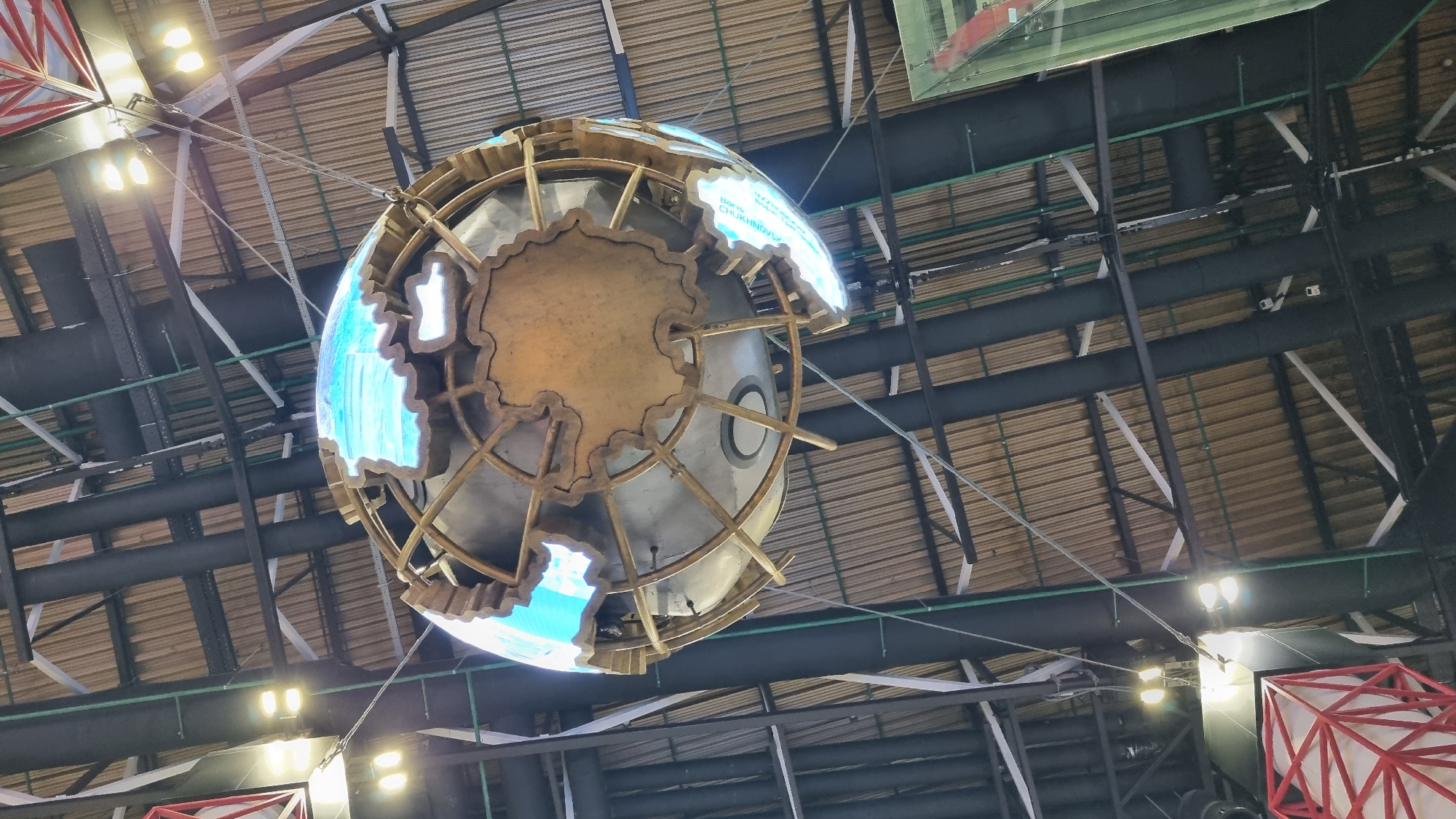 This is an upward-facing, eye-level, medium shot of a large, stylized globe sculpture hanging from the ceiling of a building. The globe features a metallic, golden-colored framework with cutouts shaped like continents. The continents are illuminated from within with a bright, electric blue light. The interior of the globe appears to be a metallic sphere with a central circular element. The sculpture is suspended by wires and cables from a complex ceiling structure composed of dark, industrial-looking beams and corrugated, tan-colored roofing material. Multiple black pipes and white support beams are visible in the background. Several light fixtures are mounted on the ceiling, emitting a bright yellow light. In the lower left corner, a red geometric sculpture with white internal elements is partially visible. To the right, a portion of a green glass structure is also present. The overall environment suggests an indoor public space, potentially a museum, exhibition hall, or transportation hub. The text "BOYS CHUKN...V..." is partially visible on the side of the globe. The scene is static and does not depict any people or discernible action. The location is identified as Khimki, Russia.