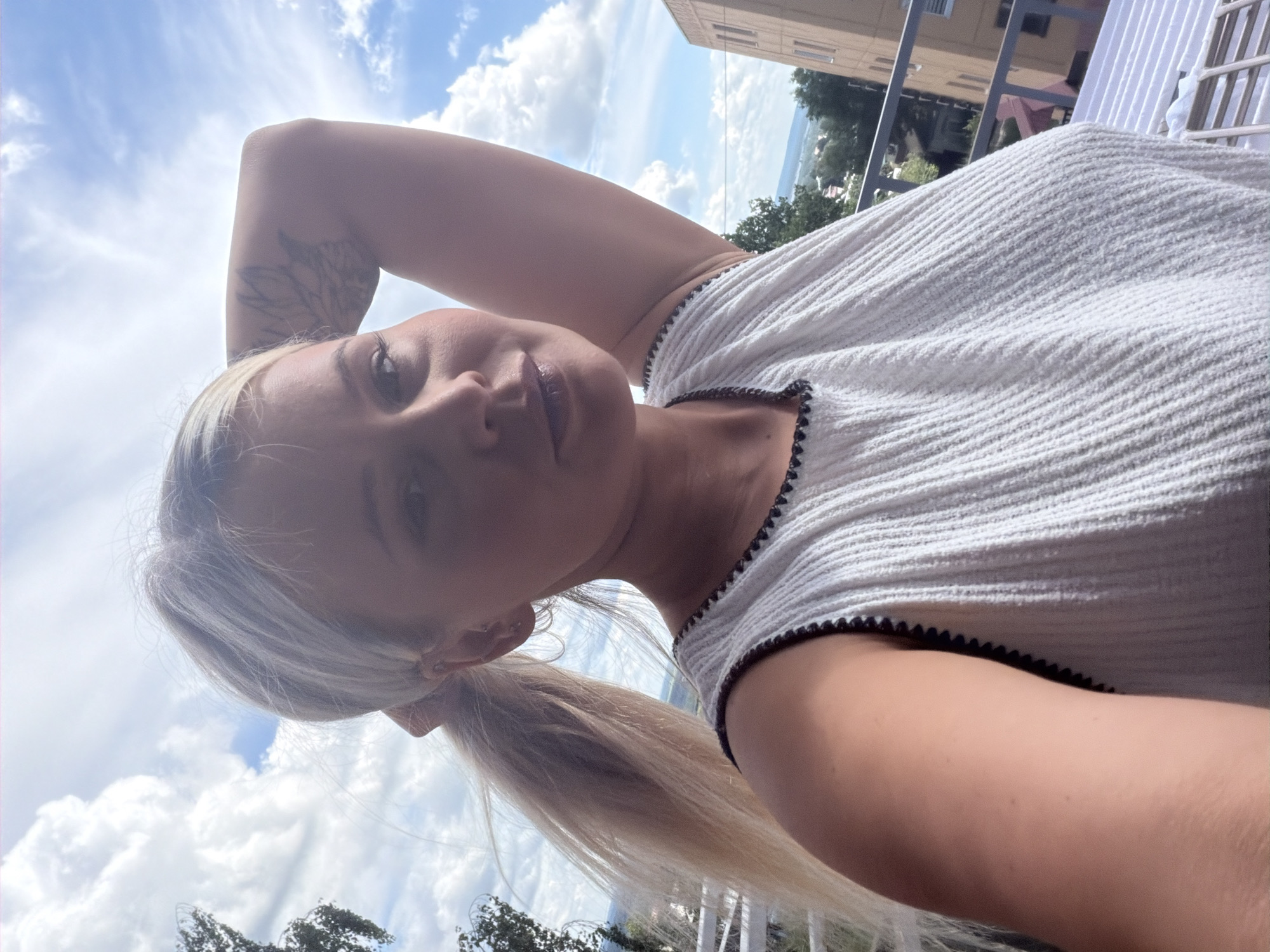 This is a portrait of a young woman outdoors, possibly on a balcony or patio, with a cloudy sky in the background. She has blonde hair styled in a ponytail and is wearing a white textured top with a dark trim. Her arm is raised behind her head, and she is looking towards the camera. The lighting suggests it is daytime, with bright sunlight filtering through the clouds. There are buildings and some greenery visible in the background, indicating an urban or suburban setting. The image appears to be a selfie, with the camera angled slightly from above. The woman's expression is neutral or slightly contemplative. There is no visible text in the image. The provided location context of Sarapul, Russia, places this scene within that geographical area.