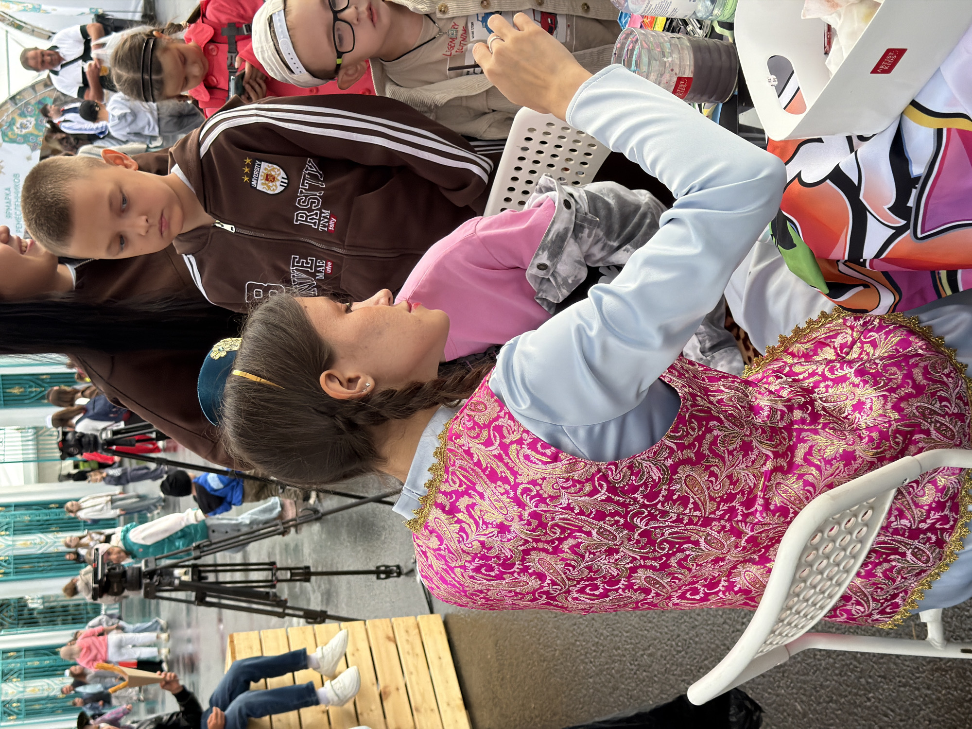 In an outdoor urban setting, a young woman with braided dark hair, wearing a light blue long-sleeved shirt and a pink vest with gold embroidery, is seated and engaged in an activity involving art supplies. She is positioned in the foreground, facing left and slightly away from the camera, with her right hand extended. In front of her, on a table or surface, are various art supplies including a white plastic container with colorful designs, a glass filled with dark liquid, and a water bottle. A white plastic chair is visible to her left.
Behind her, several people are present, suggesting a public event or gathering. A young boy in a brown hooded sweatshirt is looking down. Another child, a girl in a red jacket, is also visible in the background. In the mid-ground, two tripods with cameras are set up. Further back, other individuals and a building with decorative turquoise elements can be seen. The ground appears to be wet, indicating recent rain. The overall atmosphere suggests a lively public space, possibly a fair or festival, where artistic activities are taking place.
