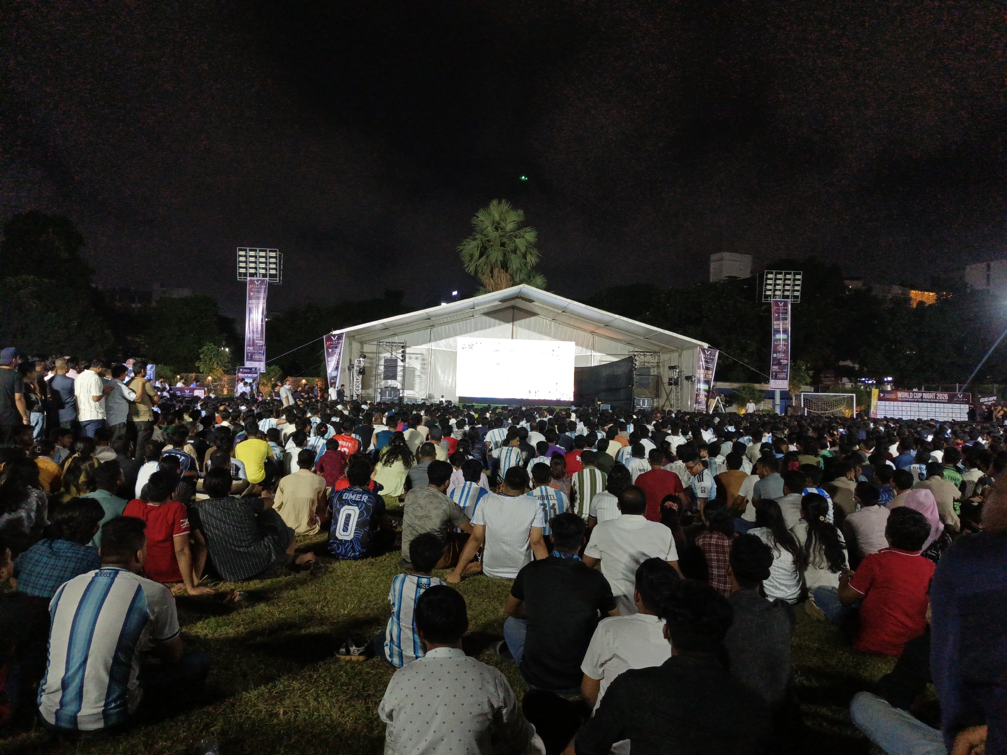 A large, outdoor crowd gathers on a grassy field at night to watch a projection on a large screen, possibly a sporting event. The scene is lit by floodlights positioned on tall poles and lights from a white tent structure in the background. Many people are seated on the ground, facing the screen, with some standing in the back. The atmosphere appears to be one of communal viewing. The location is identified as Dhaka, Bangladesh, with specific mention of the Bashundhara Sports Complex as a venue. The overall impression is a lively public event with a focus on shared entertainment.