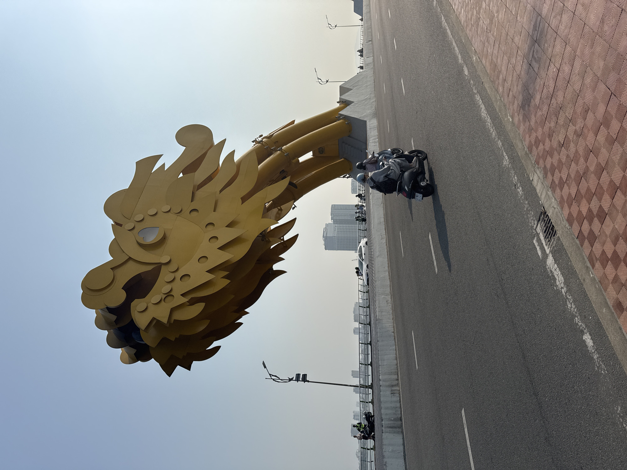 A low-angle, outdoor shot depicts the head of a large, golden dragon sculpture positioned above a multi-lane highway, likely the Dragon Bridge in Đà Nẵng, Vietnam. The dragon's head, elaborate in design, features stylized scales, whiskers, and horns. The highway below is divided by white lane markings, and several motorbikes are present on the road, carrying riders and passengers. The city skyline is visible in the hazy background, featuring tall buildings. Sidewalks on either side of the road are brick. Street lights stand tall beside the road. The sky is pale blue, suggesting a time of day with ample sunlight.