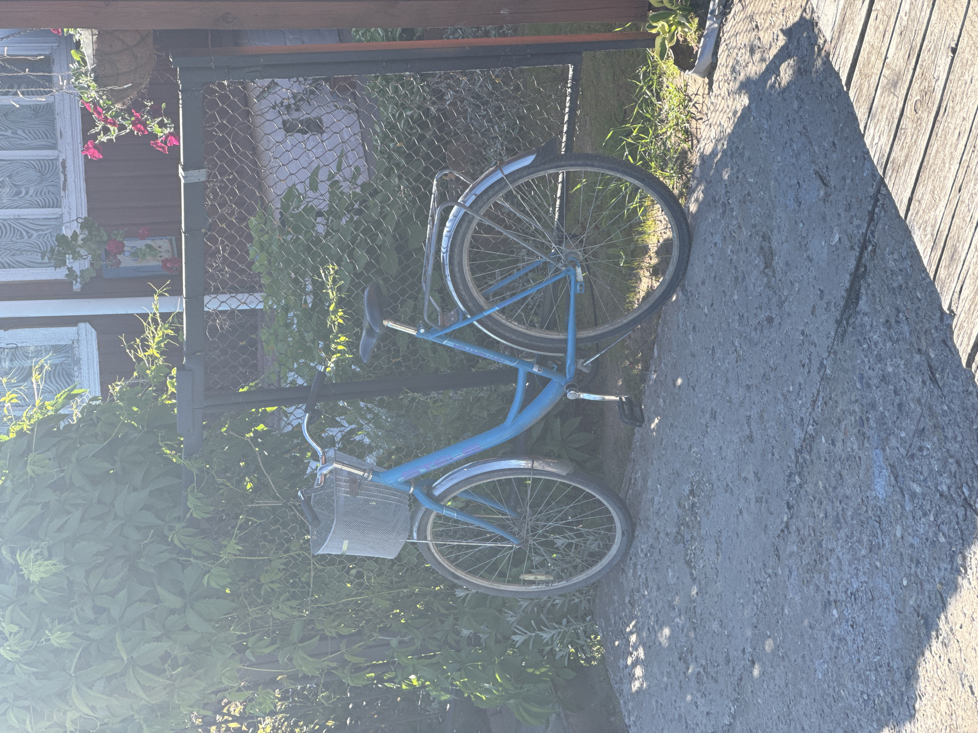 A blue bicycle with a wire basket is parked on a concrete path next to a chain-link fence and lush green foliage. The bicycle has a kickstand engaged, suggesting it is stationary. In the background, a wooden building with windows is visible, along with hanging flower baskets. The scene appears to be outdoors on a sunny day, casting dappled shadows on the ground. The location is identified as Ponaryevo, Russia, likely a residential area given the building and garden elements. No people are present in the image.
