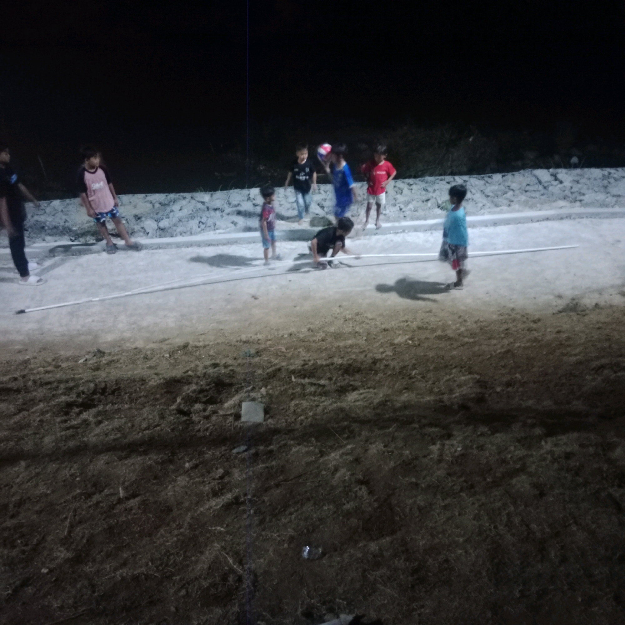 An undated, low-light, medium-angle outdoor photograph taken at night shows a group of children playing on a dusty, uneven surface. The background is dark, suggesting nighttime, with a dark, rough textured wall or embankment behind the children. In the foreground, the ground is covered in dirt and sparse vegetation.
Several children are visible, some actively engaged in play while others stand or observe. A child in a black shirt and light blue shorts is kneeling and appears to be interacting with a white pole lying on the ground. Another child, wearing a blue shirt and shorts, stands to the right of the kneeling child. To the left, a child in a pink and black shirt and patterned shorts stands with their back to the camera. Further to the left, a child in a red shirt and white shorts stands facing the group. In the middle ground, a child in a blue shirt and red shorts is partially obscured and appears to be holding a ball. Another child in a black t-shirt with a graphic print and light-colored pants is visible to the left. In the far left of the frame, a child in a dark t-shirt and black pants is standing, facing away from the camera.
The environment appears to be an undeveloped or recreational area, possibly a field or park, in the city of Tlogorejo, Indonesia. The presence of a white pole suggests some form of informal game or activity is taking place. The lighting is dim, casting shadows and creating a slightly grainy appearance.