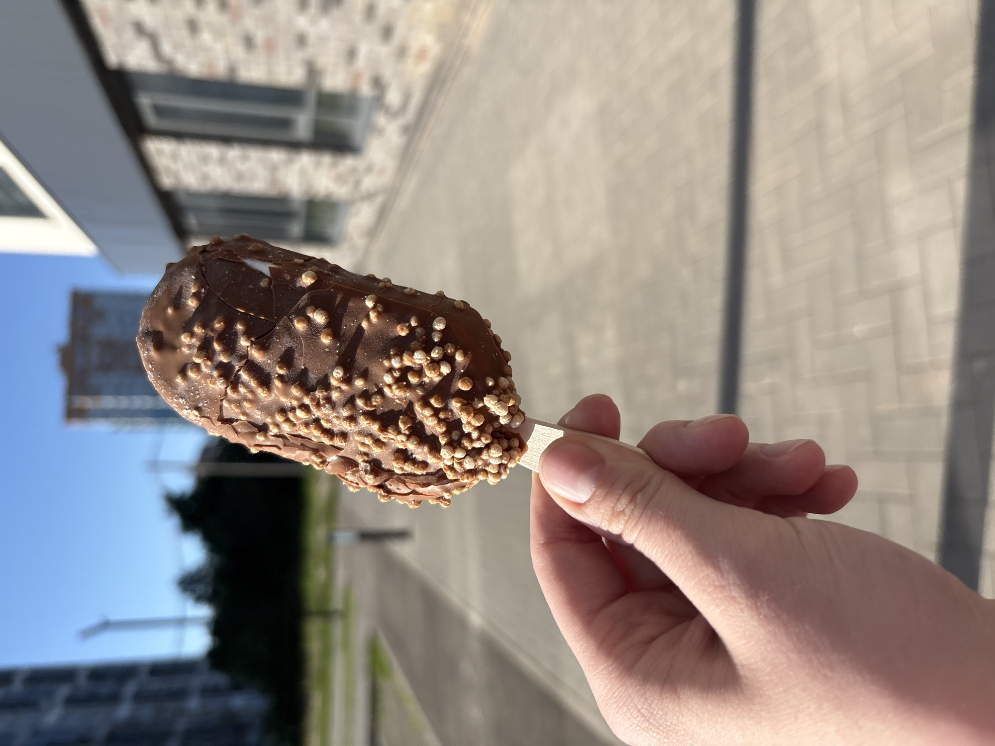 A person's hand, with light skin and short fingernails, holds a chocolate-covered ice cream bar on a wooden stick. The ice cream bar is partially submerged in a cracking chocolate shell adorned with small, spherical, light brown crisped rice pieces. In the background, a paved walkway with a light gray, rectangular pattern stretches out towards a blurred urban landscape under a clear blue sky. To the left, a dark silhouette of trees and tall, indistinct buildings are visible. To the right, a modern building with a brick facade and large windows is partially in frame. The scene appears to be an outdoor setting in a city.