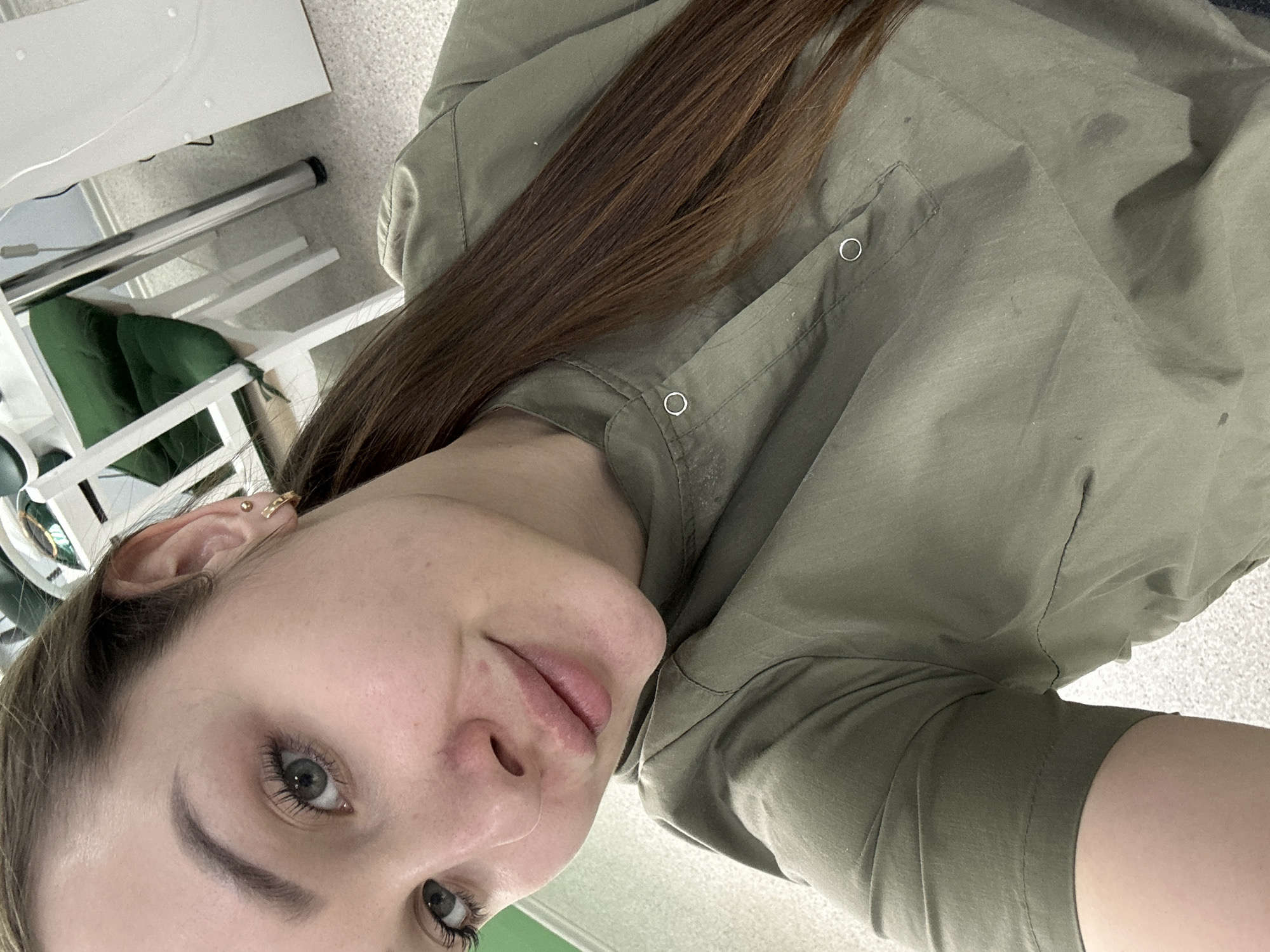 A selfie taken at an oblique angle shows a fair-skinned woman with shoulder-length brown hair, wearing a green button-up shirt. She has gray eyes and light eyebrows. A gold hoop earring is visible in her left ear. The background includes a white shelving unit with green fabric, and what appears to be a carpeted floor. Location information (Chaikovsky, Russia) is not discernible from the image.