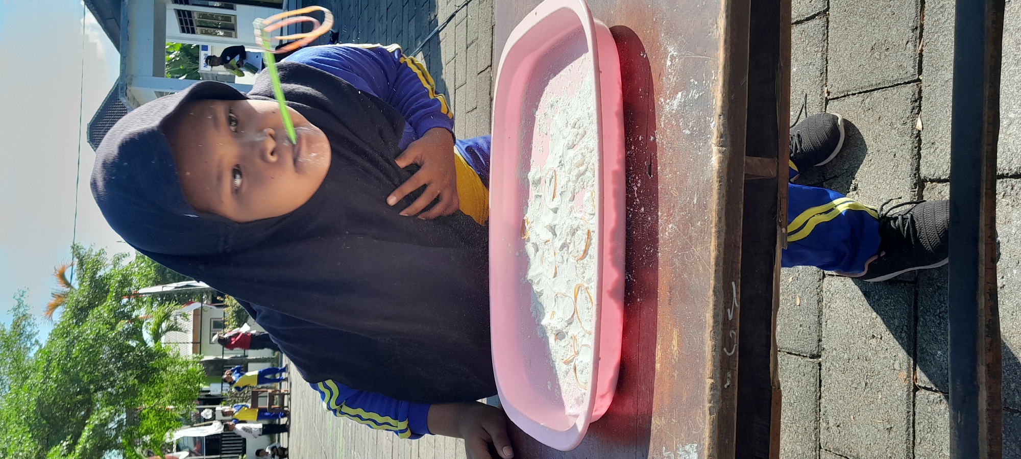 In Rogojampi, Indonesia, a young person wearing a black hijab and a blue and yellow athletic uniform is the primary subject of this outdoor photograph. They are actively participating in a game, as evidenced by the green straw held to their mouth, with bubbles or mist being blown out, and a pink plastic tray positioned on a wooden table in front of them. The tray contains a white powdery substance, possibly flour, with several small, circular objects, likely rubber bands, scattered within it. The background suggests an open area with buildings, trees, and other people, indicating a public or school event. The overall lighting suggests it is daytime with sunlight casting shadows.