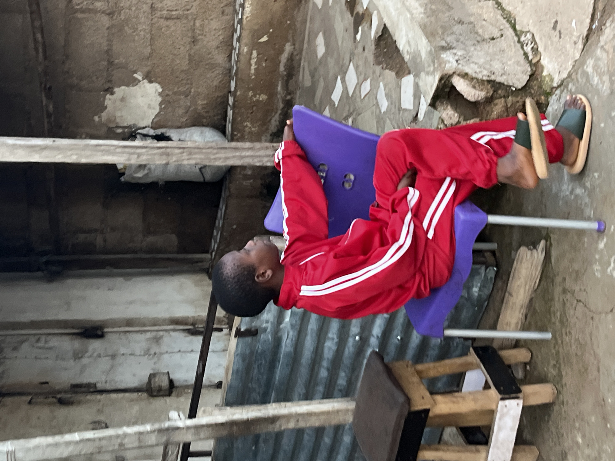 In Jos, Nigeria, a young person wearing a red tracksuit with white stripes is seated on a purple plastic chair outdoors. The individual is looking to the right. To the left of the chair, a brown wooden stool with a dark top is visible. The ground is a paved surface with white square tiles and some patches of dirt. In the background, a corrugated metal structure is present, and behind that, a textured concrete wall is seen. A white bag, partially ripped, is leaning against a wooden beam in the background. The scene appears to be in an urban or semi-urban environment, possibly a residential area or a marketplace.