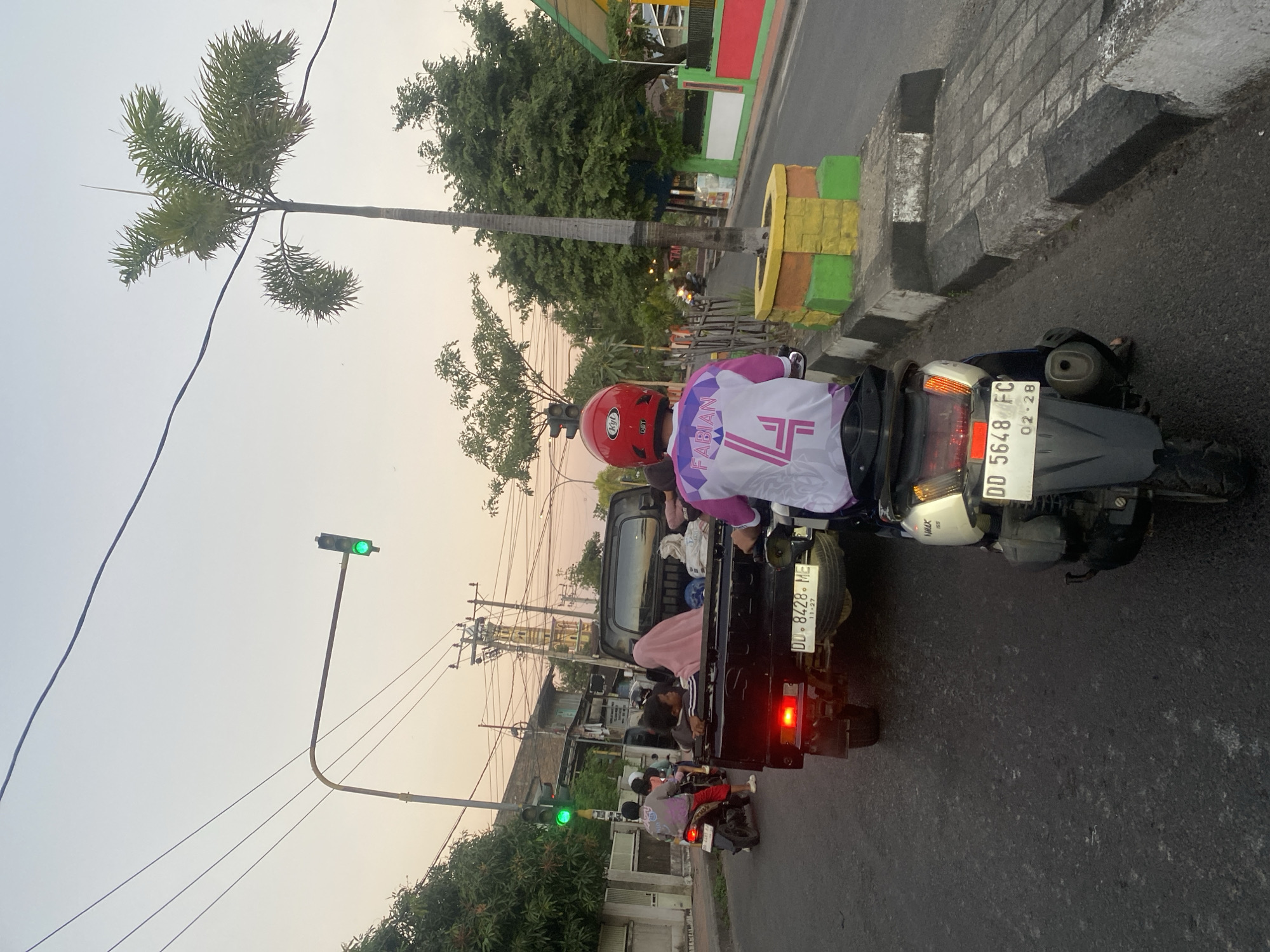 In Bantaeng, Indonesia, a view of traffic at dusk shows a black pickup truck in the foreground with several passengers, including a woman in a pink hijab. The truck's license plate reads "DD 8428 ME". A person on a scooter, wearing a red helmet and a white and purple shirt with "FABIAN 4" on the back, is positioned behind the pickup. The scooter's license plate reads "DD 5648 FC". To the left, a motorcycle with a rider wearing a pink and gray shirt and red pants is visible. A traffic light with a green signal is positioned above the intersection. Palm trees and other foliage line the street, and utility poles and wires are present. The sky is a pale orange, indicating sunset.
