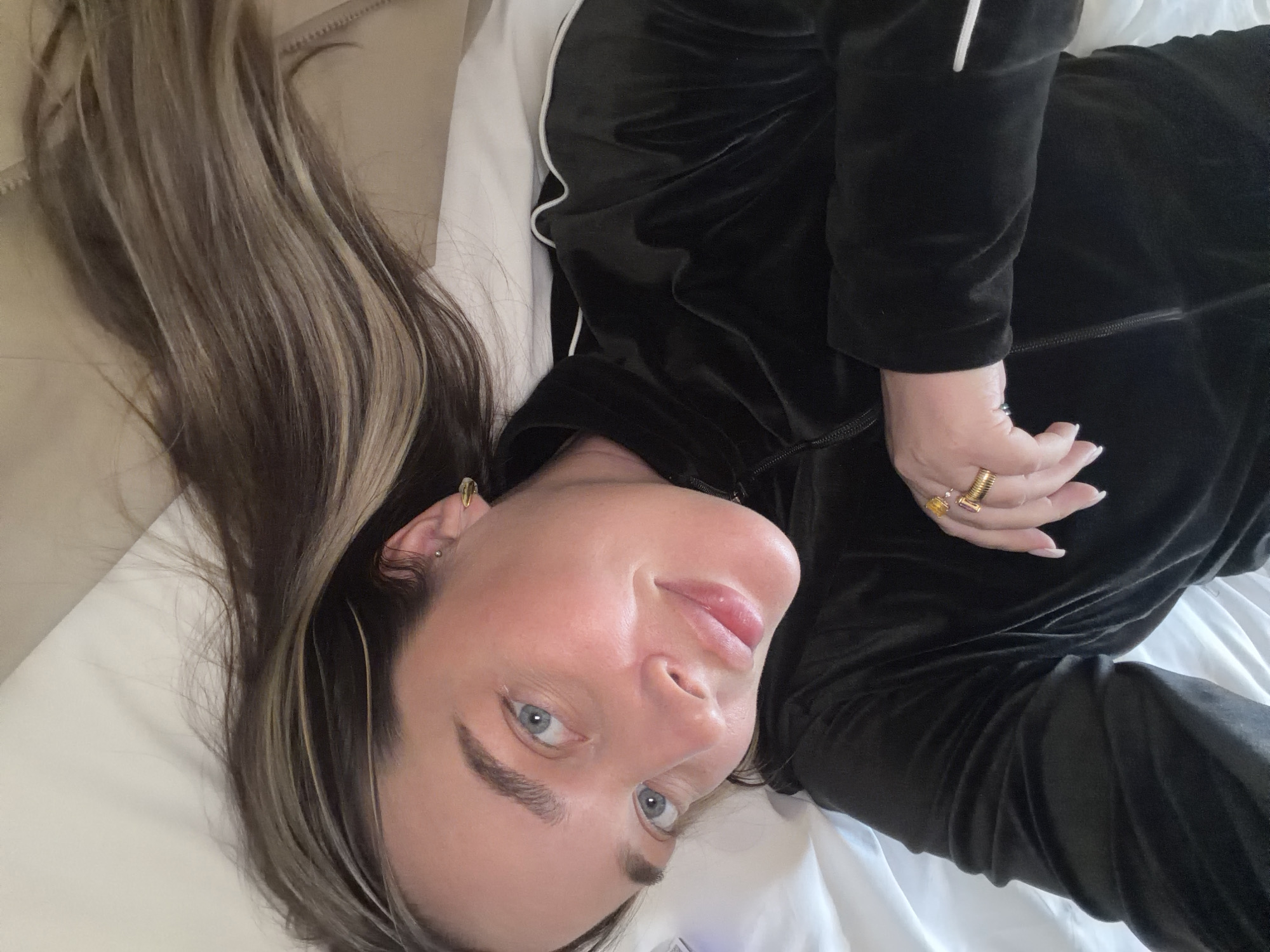 A woman with long, highlighted brown hair lies on a white bed. She has blue eyes and wears gold earrings and rings. She is wearing a black velvet tracksuit with white stripes down the sides. The scene does not appear to be in Moscow, Russia. There is no identifiable landmark or cultural reference that would place the image in that location.