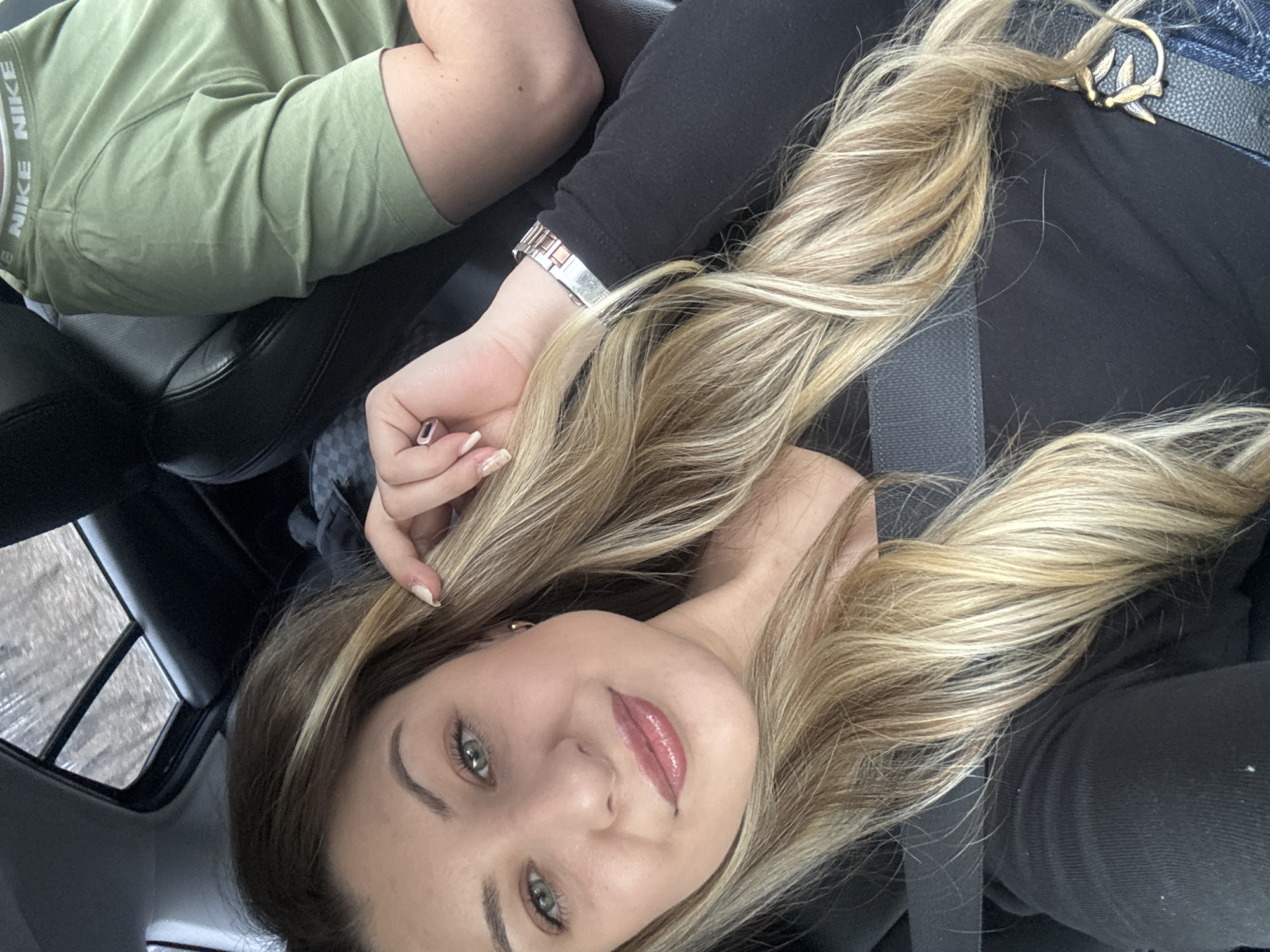 A selfie photo taken inside a car, presumably in motion given the out-of-focus scene visible through the sunroof, shows a woman with long, blonde hair, fair skin, and green eyes. She is wearing a black long-sleeved shirt, and the seatbelt is fastened across her body. On her wrist is a silver watch. A man's arm and torso, clad in a green Nike t-shirt, are visible in the upper left corner. The location in Goritsa, Bulgaria is undetermined from the available visual information.