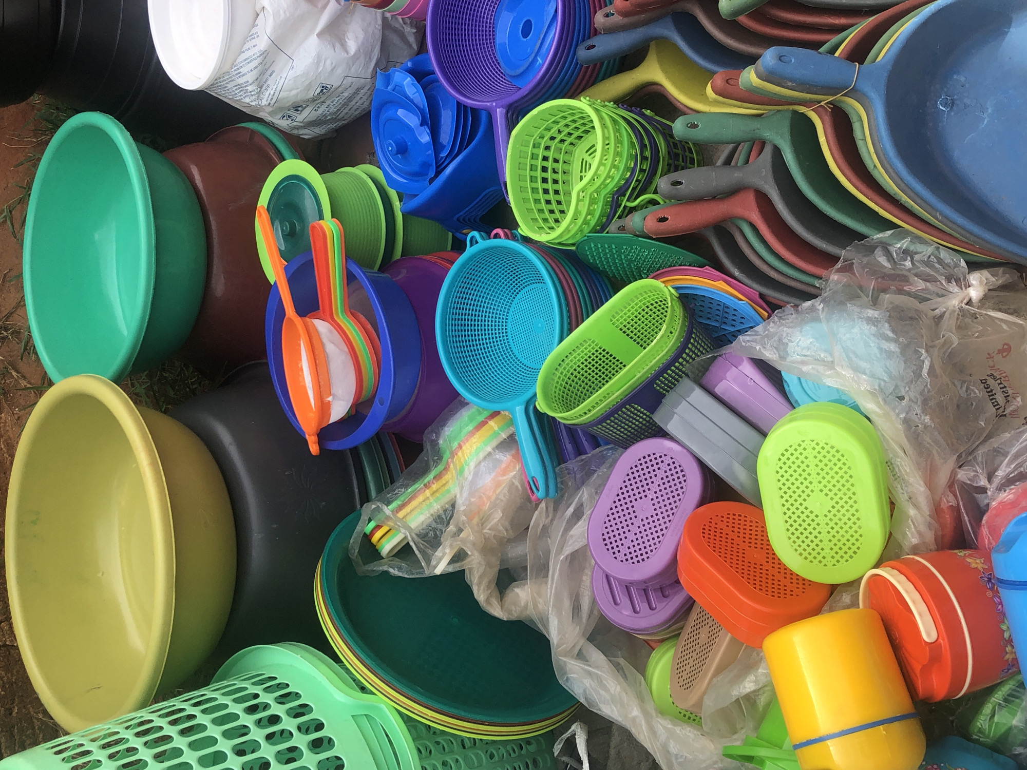 In Amaeke Abam, Nigeria, an eye-level shot reveals a jumbled pile of colorful, plastic household items outdoors, including bowls, dustpans, strainers, and buckets in varying sizes and shapes. The items are of various colors, including green, yellow, orange, red, blue, and purple. A white plastic bag is also visible in the pile. The scene is set on the ground, possibly dirt or grass.