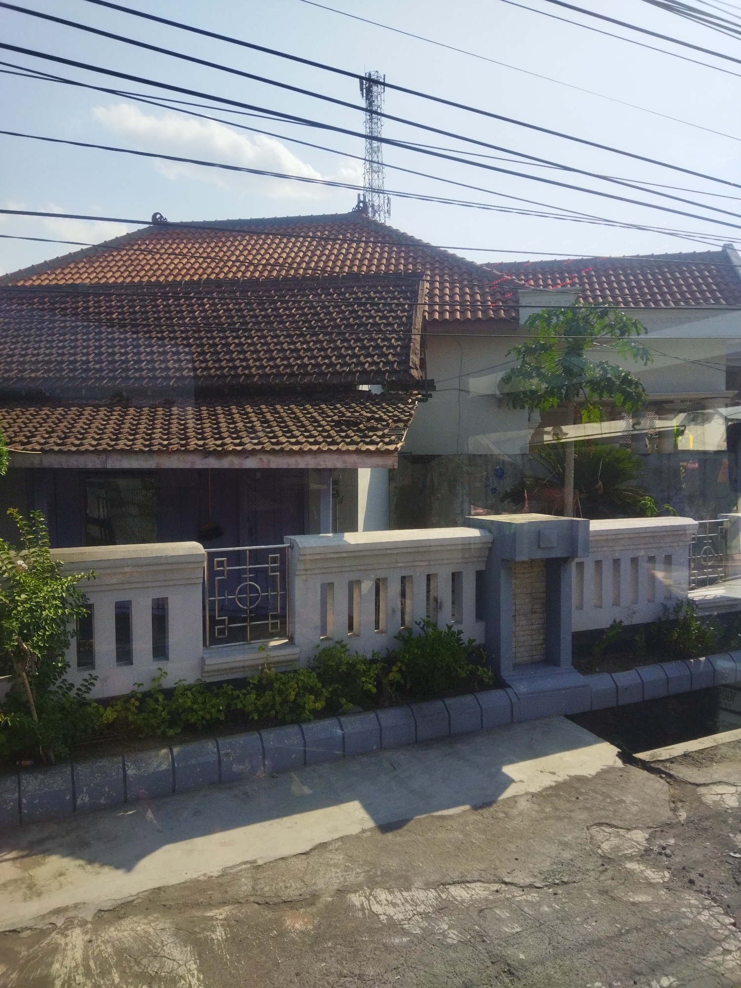 The image shows a residential street scene in Begajah, Indonesia, dominated by a house with a tiled roof. Overhead, a complex network of power lines stretches across the frame, partially obscuring a tall telecommunications tower in the background. In the foreground, a low, modern fence separates the property from the street. Green bushes and small trees are planted along the fence line. The street itself is paved with concrete, showing signs of wear and cracking. The sky above is clear blue with a few wispy clouds, indicating daylight.