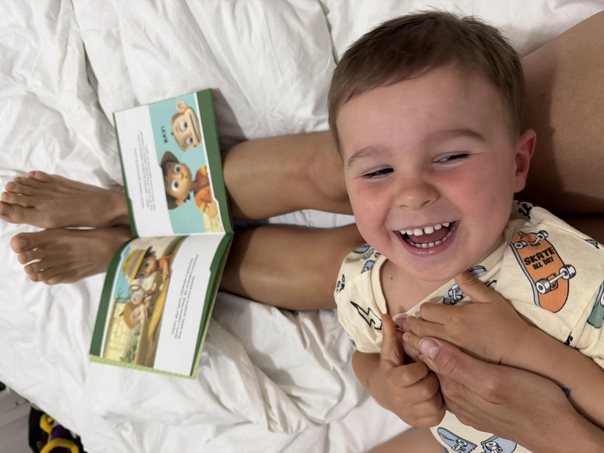 A small child lies on a white bed, held by an adult. The child is smiling broadly, displaying his teeth. He wears a light yellow shirt printed with skateboards. An open book lies near the adult's legs. The book is decorated with cartoon characters on the front cover and an illustration inside. The adult's feet are visible near the book. The location cannot be determined as being in Budva, Montenegro based on the image.