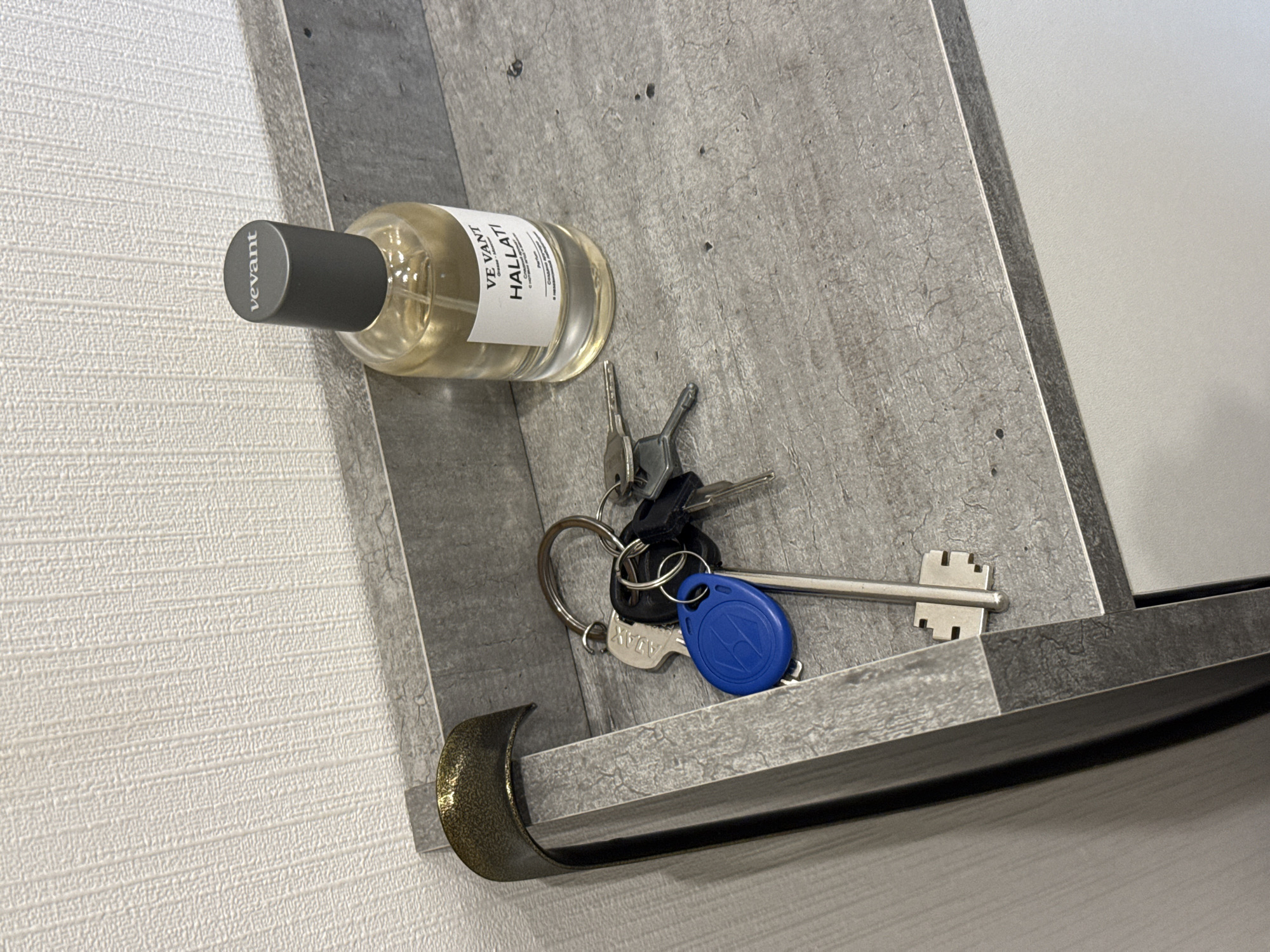 The image shows a close-up of a textured shelf with a bottle of "VE.VANT HALLATI" perfume and a set of keys. The shelf appears to be made of a grey, stone-like material with a rough texture. A curved, metallic object, possibly a hook or decorative element, is attached to the left edge of the shelf. The perfume bottle is clear glass with a grey cap and a white label. The keys are on a keyring, and one of the keys has a blue electronic access tag attached. The background is a textured white wall. The scene is indoors and the lighting suggests it is daytime. The location is specified as Lyubertsy, Russia.