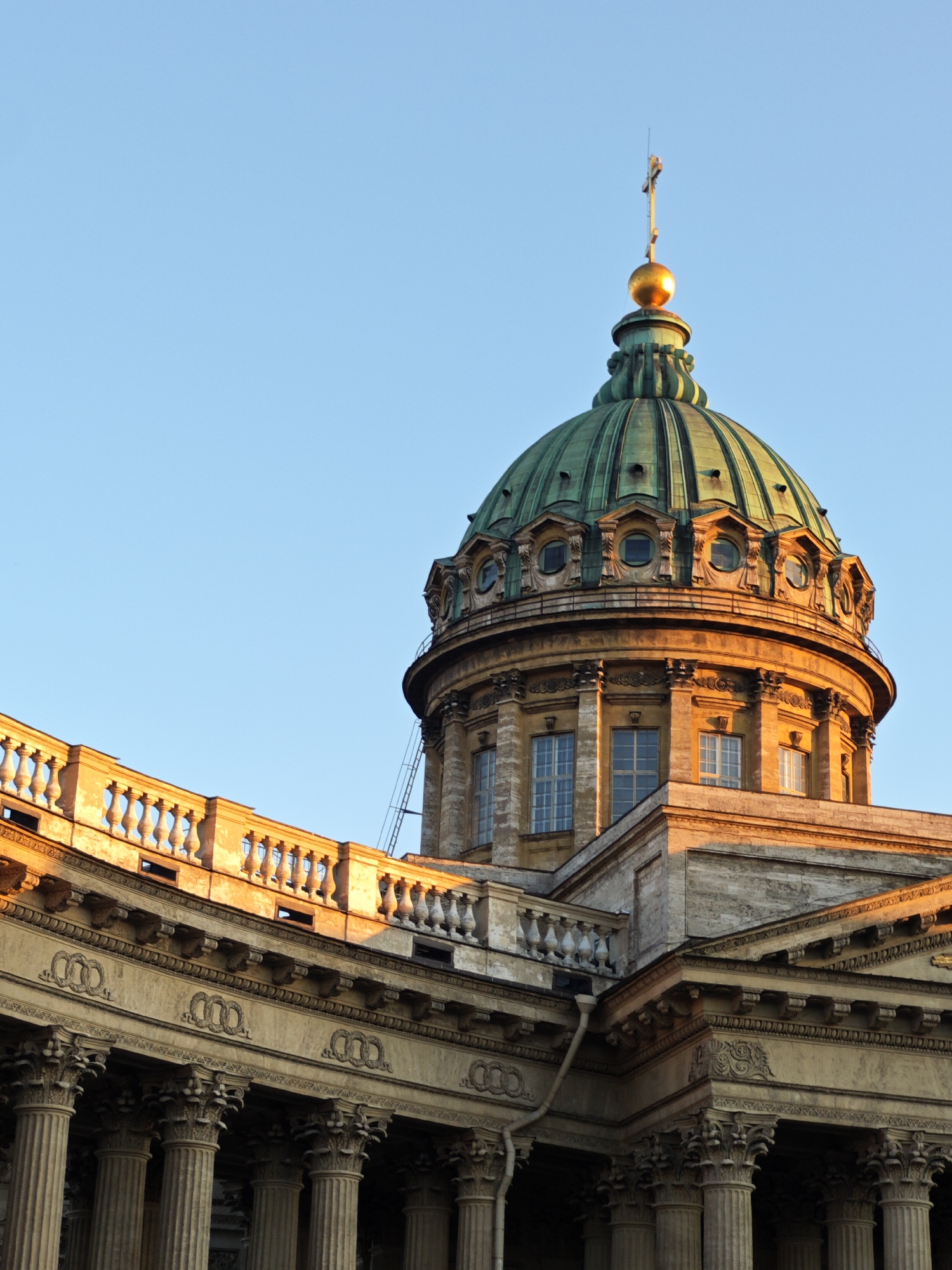 This upward-angled photograph presents a detailed view of the Kazan Cathedral in Saint Petersburg, Russia, set against a clear, light blue sky. The dominant feature is the cathedral's large, segmented dome, patinated to a verdigris green. It is topped by a lustrous golden sphere from which a slender cross extends upwards.
Below the dome, a circular drum section of the building is visible, constructed from light-colored stone, exhibiting rectangular windows framed by architectural detailing. Further down and to the left, a portion of the building's facade features a row of closely spaced Corinthian columns, supporting an entablature adorned with relief carvings, including repeating circular motifs. Above this, a balustrade with numerous stone spindles runs along the edge of a roofline.
The scene is illuminated by strong, warm sunlight, likely from early morning or late afternoon, which casts a golden hue on the right-facing surfaces and upper sections of the building, creating defined shadows that accentuate the architectural depth. A thin, grey downspout is visible running vertically on the lower left side of the structure, and a ladder is affixed to the exterior wall leading up the drum beneath the dome. No people, animals, or specific actions are depicted within the frame.