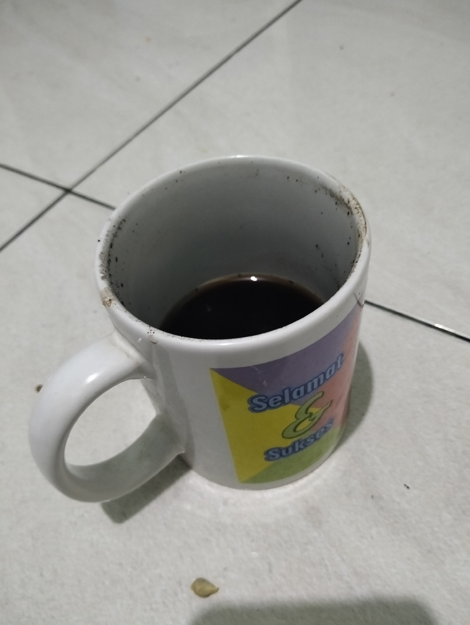 A white ceramic mug filled with dark liquid, likely coffee, sits on a tiled surface. The mug has a handle and a colorful design featuring the words "Selamat & Sukses" in stylized fonts. The rim of the mug appears to have some dried residue. The tiled floor is light-colored with visible grout lines. The overall lighting suggests an indoor environment, potentially in Paciran, Indonesia, though there are no explicit visual cues to confirm the location.