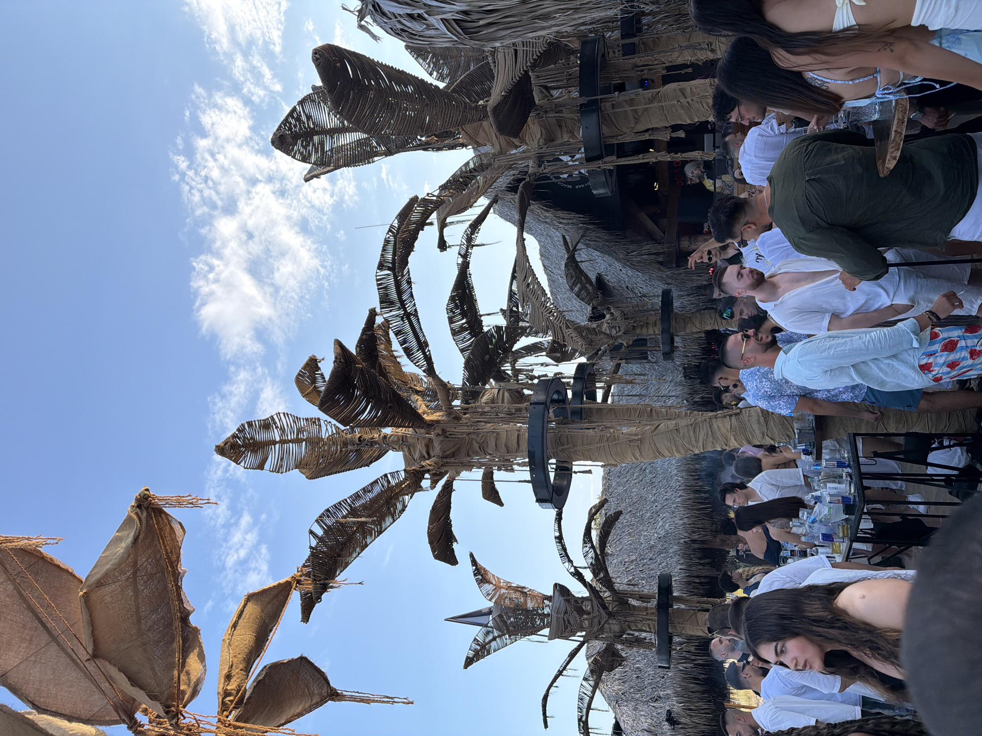 The image captures a lively outdoor gathering, possibly a beach bar or club, set against a bright blue sky with scattered clouds. The scene is dominated by stylized, artificial palm trees constructed from rope and woven materials, creating a tropical ambiance. A thatched roof structure, likely part of the establishment, is visible in the background.
A diverse crowd of people, dressed in casual summer attire, are engaged in various activities. Many are standing and conversing in groups, while others appear to be ordering or consuming drinks from tables laden with bottles and glasses. The atmosphere suggests a social event or party, with a relaxed and festive mood.
The location is identified as a bar in Sozopol, Bulgaria, likely on the coast given the beach-like setting and tropical decor. The time of day appears to be late afternoon or early evening, with the sunlight casting a warm glow on the scene. The image focuses on the interaction between people and the surrounding environment, highlighting the social aspect of the gathering.