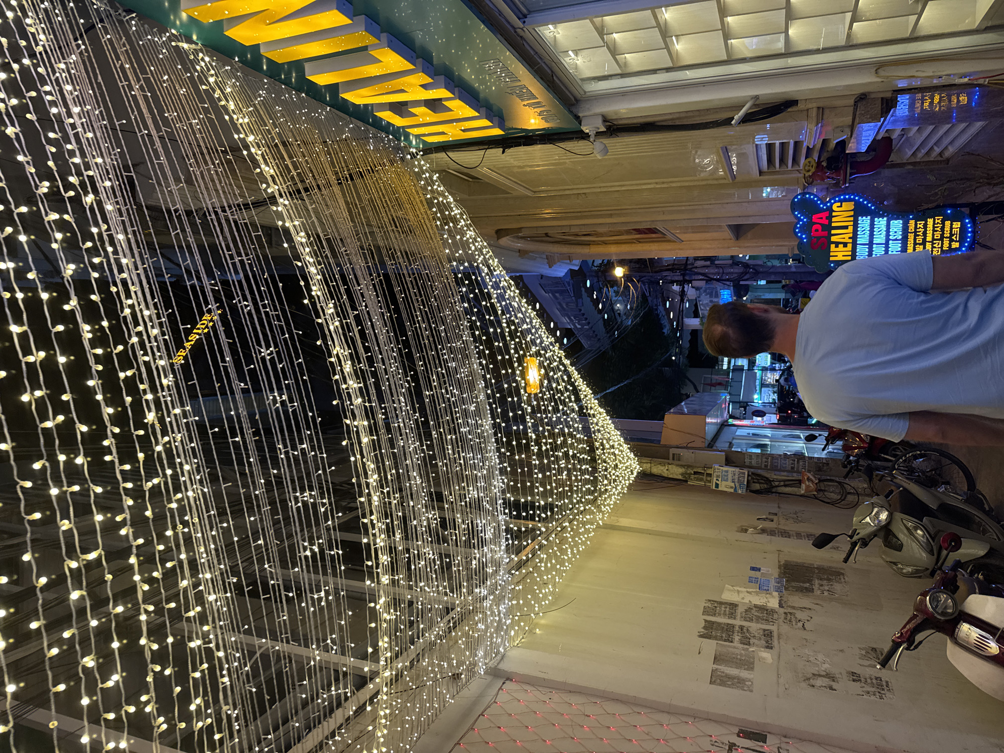 The image depicts a street scene in Unknown, Unknown, at night. A man in a light blue t-shirt is standing on the sidewalk, looking at a business or down the street. Several parked motorbikes are near him. Above, a sign with the word "HEALING" in large, vertical, blue letters with white text is visible. To the side of the man is a wall decorated with string lights, forming a curtain-like display. At the top of the frame, a yellow sign with the letters "ME CAM" is visible. The environment suggests an urban setting with businesses and possible tourist attractions. The lighting is bright and varied due to signage and streetlights.