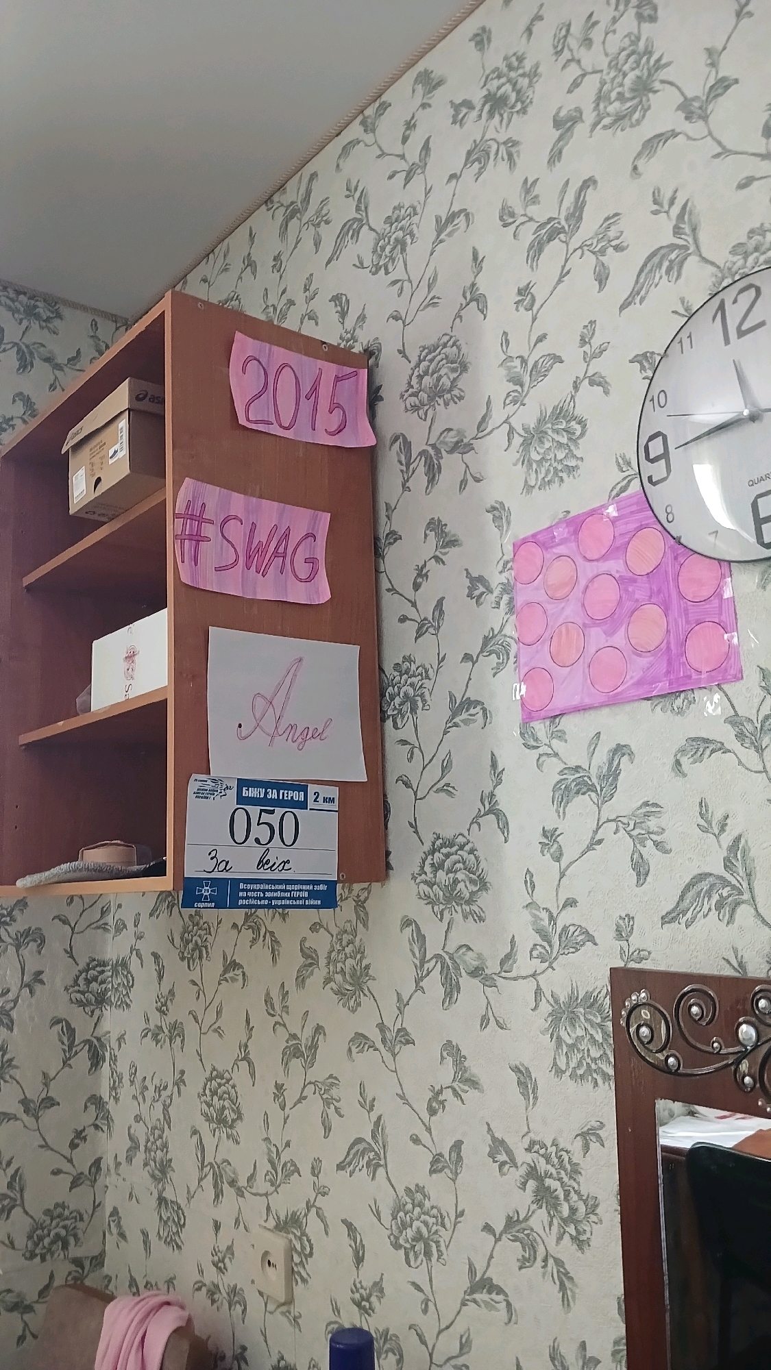 The image shows a close-up of a wall in an indoor setting, likely a room. The wall is covered with a floral wallpaper featuring a pattern of greyish-green flowers and vines on a light-colored background.
On the left side of the image, a portion of a brown wooden shelf unit is visible. It has multiple shelves. On one shelf, there is a cardboard box, possibly a shoe box, with some writing and a logo on it. On another shelf, there is a white rectangular object, possibly another box.
Attached to the front of the shelf unit are several pieces of paper. At the top, a pink piece of paper has the number "2015" written on it in purple marker. Below that, another pink piece of paper has "#SWAG" written in purple marker. Below that, a white piece of paper has the word "Angel" written in a cursive, pink font. Below the "Angel" paper, there is a rectangular item that appears to be a race bib, with text and numbers printed on it. The text includes "БІЖУ ЗА ГЕРОЯ" (Running for heroes) and "2 км" (2 km), along with the number "050" and "За всіх" (For all).
To the right of the shelf unit, a large analog clock is mounted on the wall. It has a white face with black numbers and hands, and the brand name "QUARTZ" is visible near the bottom.
Further to the right, a piece of paper with a pink background and several purple circles drawn on it is attached to the wall.
The bottom right corner of the image shows a part of a wooden frame with decorative carvings, suggesting a mirror or a picture frame. A part of a white object is visible within this frame.
There are no people visible in the image. The overall impression is of a decorated wall space with personal items and a clock.
The request to provide the location in the city of Haivoron, Ukraine is not possible based on the visual information present in the image. The image itself does not contain any identifiable landmarks or clues that would allow for such a precise localization.