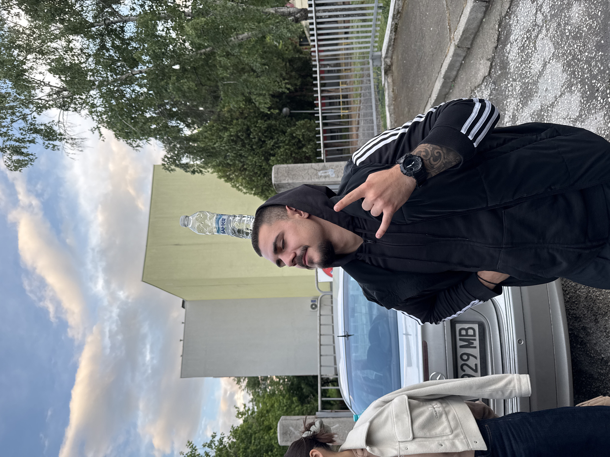 A man balances a water bottle on his head while standing in front of a silver Mercedes-Benz in Пловдив, Bulgaria. The man is wearing a black Adidas tracksuit and a watch on his left wrist, which is adorned with a tattoo. He is making a "rock on" hand gesture. A woman wearing a white jacket and jeans is standing in front of the car. The background shows a building, trees, a fence, and a cloudy sky.