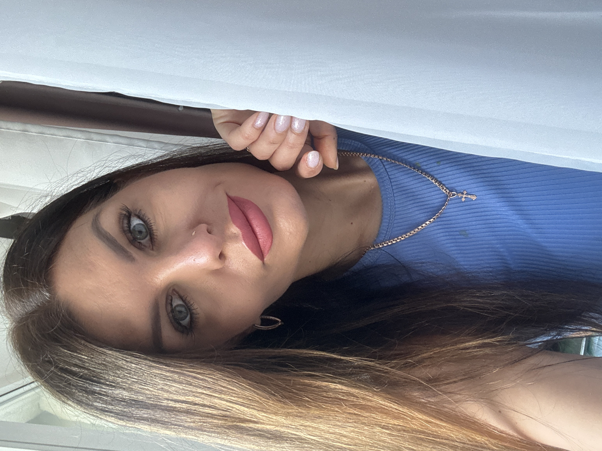 A woman with long, ombré brown and blonde hair is partially visible in a close-up, head-and-shoulders shot. She is wearing a light blue ribbed top and a gold chain necklace with a cross pendant. Her fingernails are painted a light, sparkly pink. She is peeking from behind a white curtain. The exact location in the city of Chaikovsky, Russia cannot be determined from the provided image.