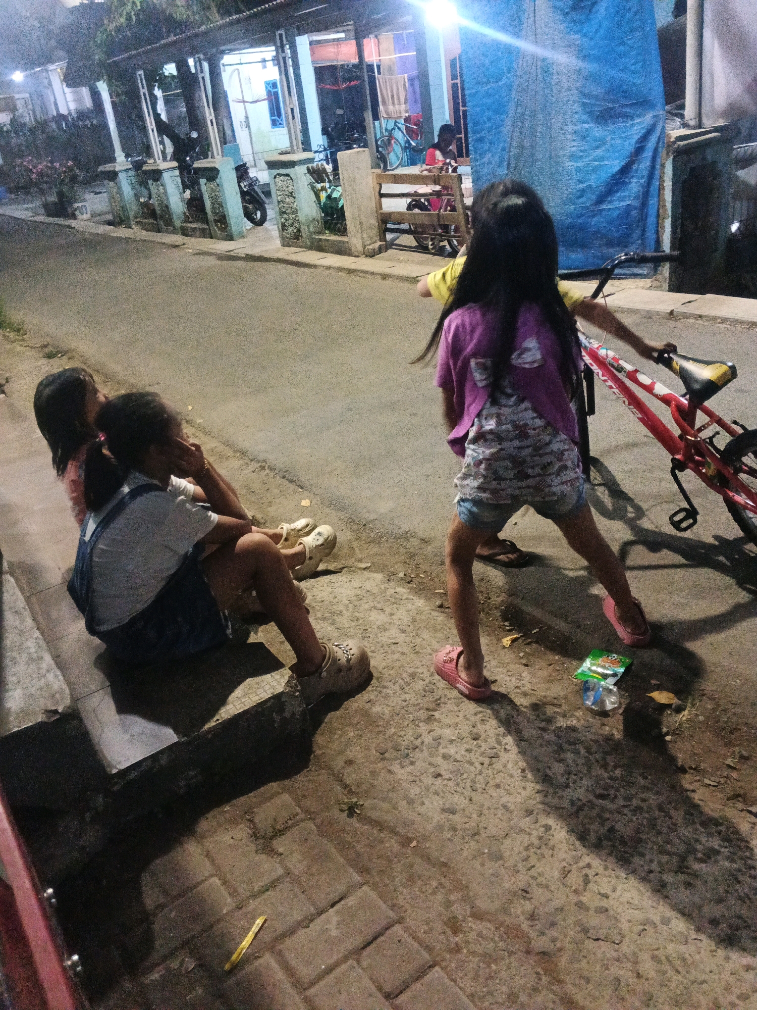 The image depicts an outdoor scene at night in Kadu, Indonesia, likely on a street or alleyway. In the foreground, two young girls are sitting on a paved surface. One girl, wearing denim overalls over a white shirt and light-colored clogs, is seated, leaning back and touching her face. The other girl, facing away from the camera, is seated behind the first, wearing a pink t-shirt and denim shorts, with her back partially against a red bicycle. A third child, also facing away from the camera and wearing a yellow shirt and patterned shorts, is standing next to the bicycle, holding its handlebars. The background reveals a building with a covered outdoor area supported by pillars, and a blue tarp draped over part of the structure. Several motorcycles are parked along the covered area. The ground is a mix of asphalt and pavers, with some debris scattered about, including a plastic wrapper and a plastic bottle. The lighting suggests it is nighttime, with artificial lights illuminating the scene.