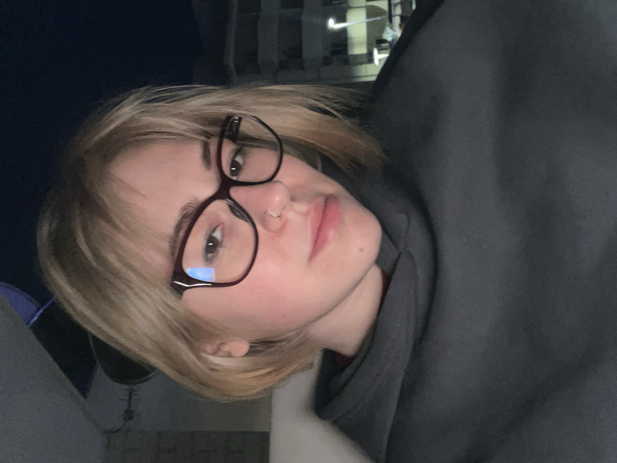A photograph captures a person with blonde hair and bangs, wearing glasses and a dark hooded sweatshirt. A nose ring is visible. The setting appears to be an indoor location, possibly a room within a building, with a blurred background that could be a city scene at night. The exact location of the city and country are not apparent in the image.