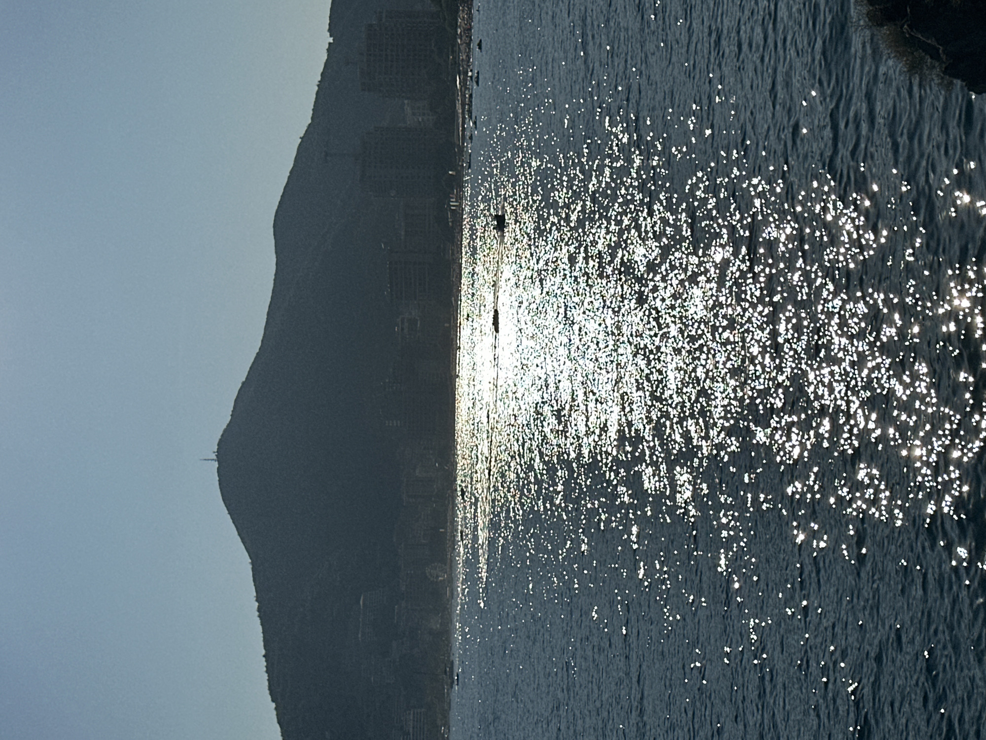 The image captures a serene coastal view of Budva, Montenegro, dominated by a silhouette of a lush, mountainous landscape against a pale blue sky. A prominent peak on the mountain range features a small antenna structure at its apex. At the base of the mountains, a cluster of buildings, appearing to be hotels or residential complexes, are visible as dark shapes against the verdant hillside. The foreground is occupied by the deep blue expanse of the Adriatic Sea, its surface dappled with the bright, shimmering reflection of sunlight. This intense sun glitter creates a luminous path across the water, broken by small, white-capped waves. In the distance, along the coastline, a few small boats are scattered on the water, some appearing to be jet skis, suggesting recreational activity. The overall impression is one of a tranquil, sun-drenched day by the sea, with the natural beauty of the landscape meeting the urban development of Budva.
