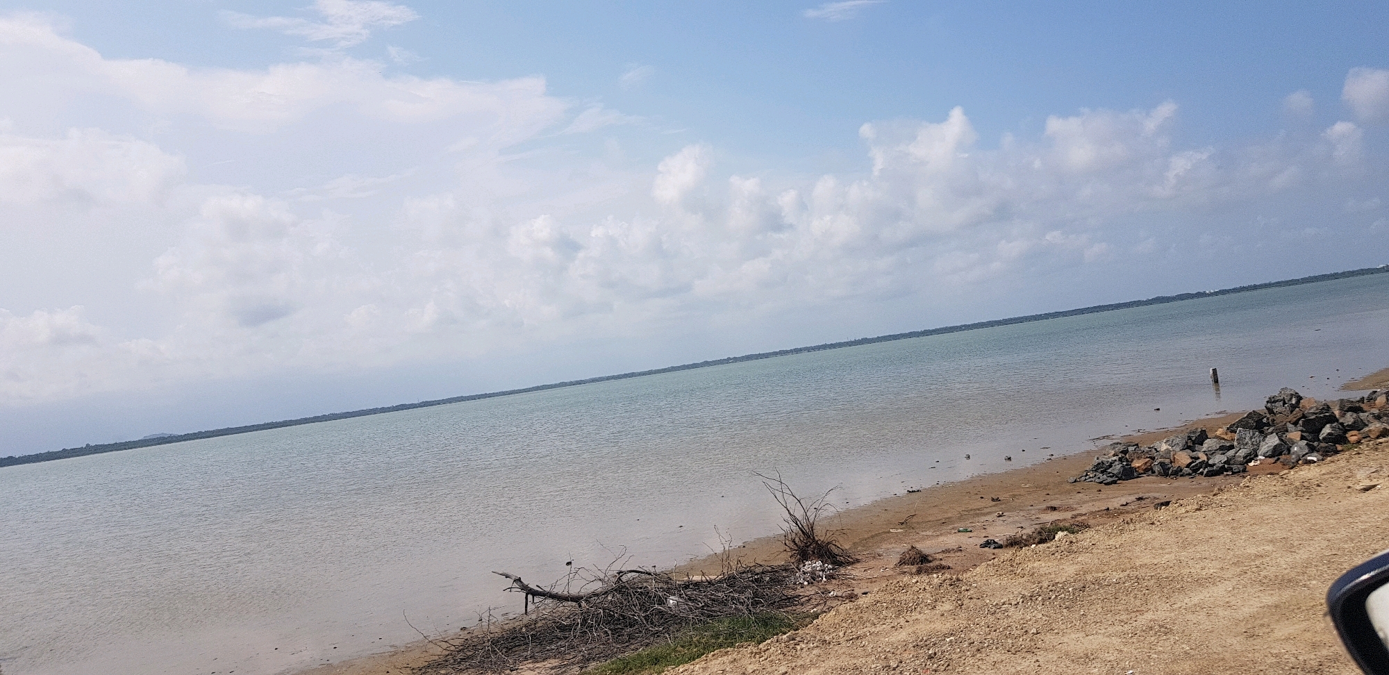 A wide-angle, slightly angled landscape shot captures a body of water under a partly cloudy sky. The water, a muted blue-green, fills the majority of the frame, meeting a sandy shoreline. On the shoreline, debris including branches, plastic waste, and a small pile of rocks are visible. The land appears dry and sandy, with sparse vegetation. In the far distance, a thin line of green vegetation suggests land. The sky is a pale blue with scattered, puffy white clouds. The scene is serene and appears to be a coastal or large lake area.