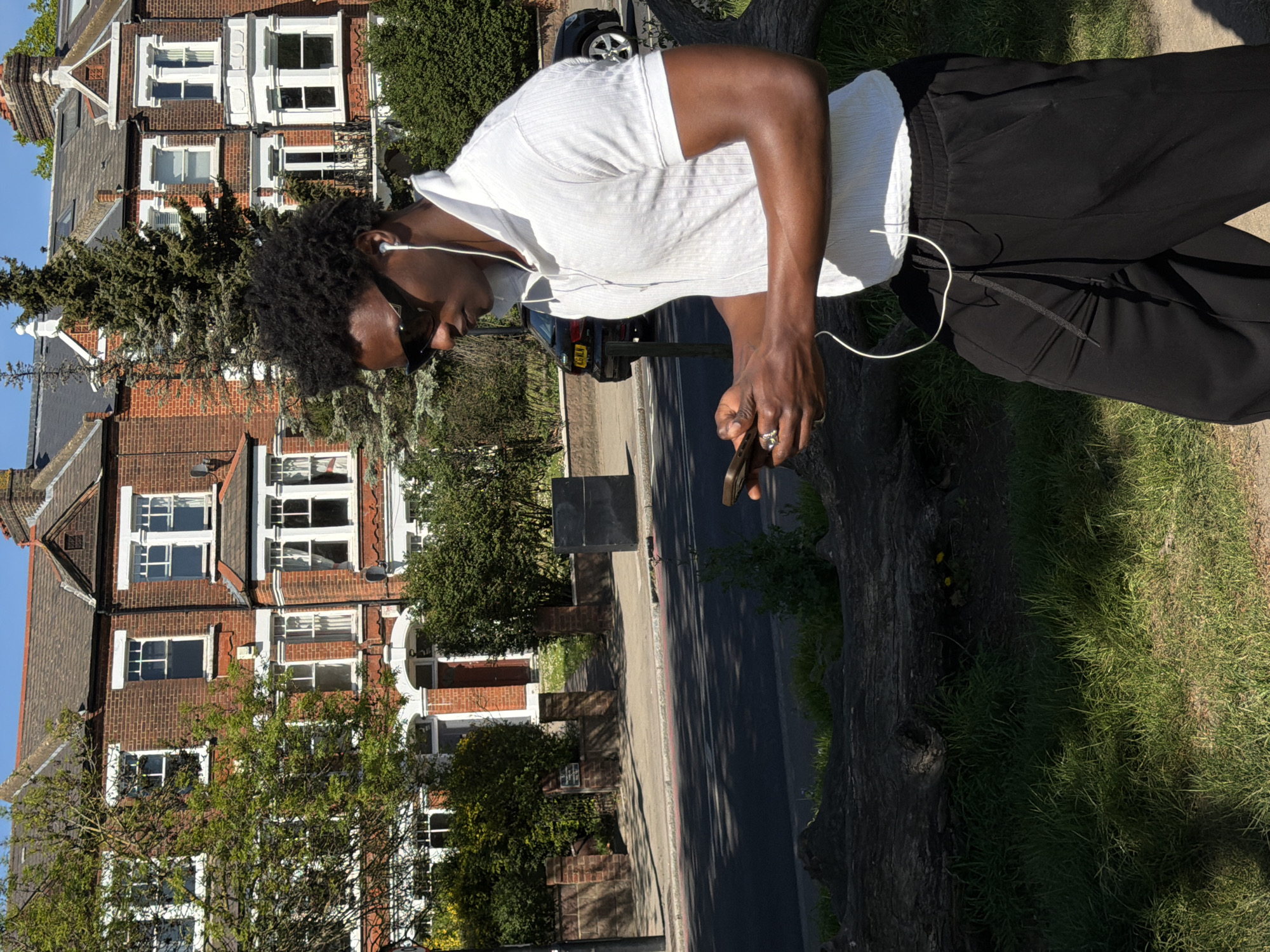 A man stands on a grassy verge in what appears to be a residential area of London, United Kingdom. He is facing away from the camera with his head angled downwards and looking towards his phone. He is wearing a light-colored short-sleeved shirt, dark trousers, and in-ear headphones. The man's hair is dark and appears to be a natural afro style. He is holding a smartphone in his right hand. He is wearing sunglasses. In the background, there are rows of brick houses with white-framed windows. Some houses are partially obscured by trees. A road with a parked car is visible between the houses and the grassy verge where the man is standing. The light suggests it is a sunny day.