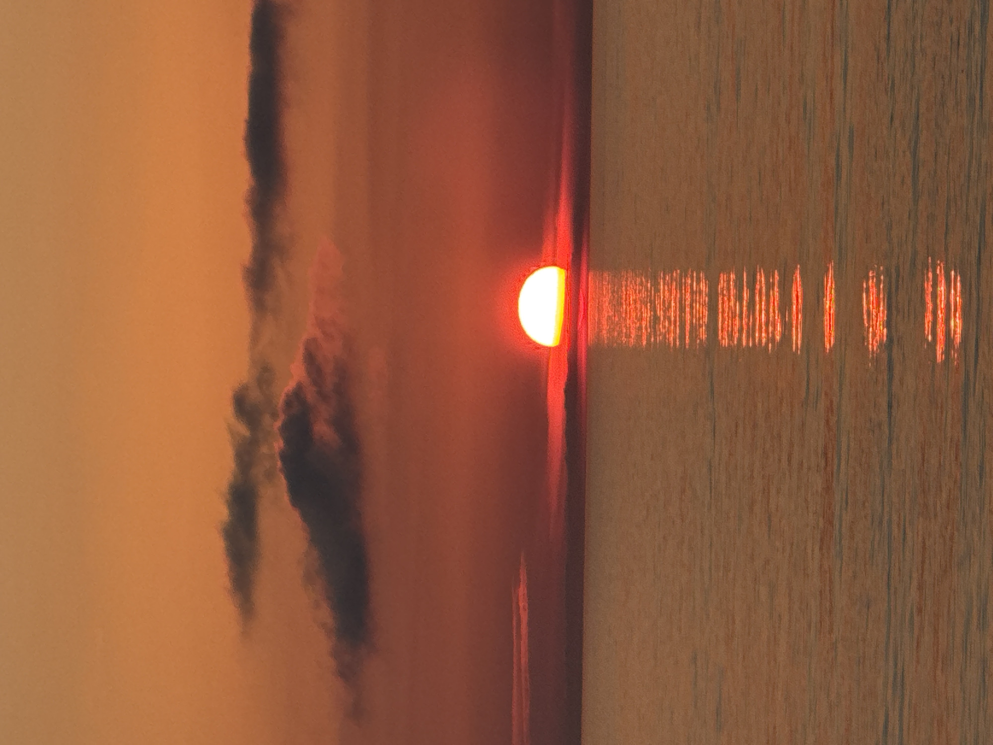 The image is a landscape photograph taken at sunset, most likely near a body of water. The sun is partially obscured by clouds, creating a half-circle effect with a bright, orange-yellow glow. A line of light reflects off the water, appearing as a series of dashes, diminishing with distance. To the left of the sun, dark, blurred clouds are visible against a pale orange sky. The scene evokes a sense of calm and serenity. The image does not depict the city of Batumi, Georgia.