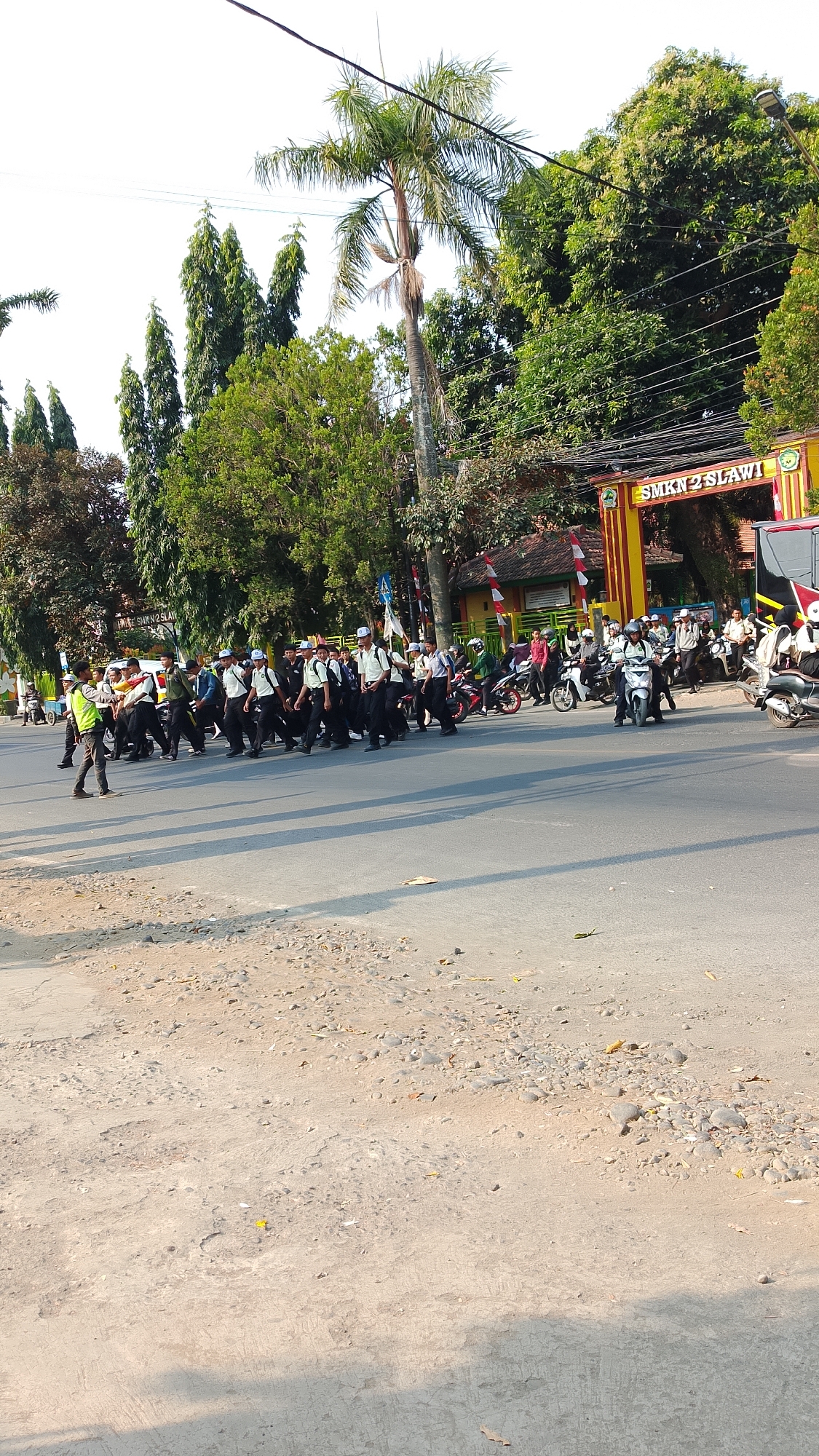 The image depicts a street scene in front of "SMKN 2 SLAWI" in Procot, Indonesia. A group of students, all wearing white shirts, dark pants, and white hats, are walking across the street. Some are wearing backpacks. One student in a yellow vest appears to be guiding the group. Several motorcycles are present, with some students riding them and others parked. The school building is visible behind a gate with the school's name prominently displayed. Trees and palm trees line the street, and power lines are strung overhead. The road is paved, with some gravel and debris on the side. The lighting suggests it is daytime.