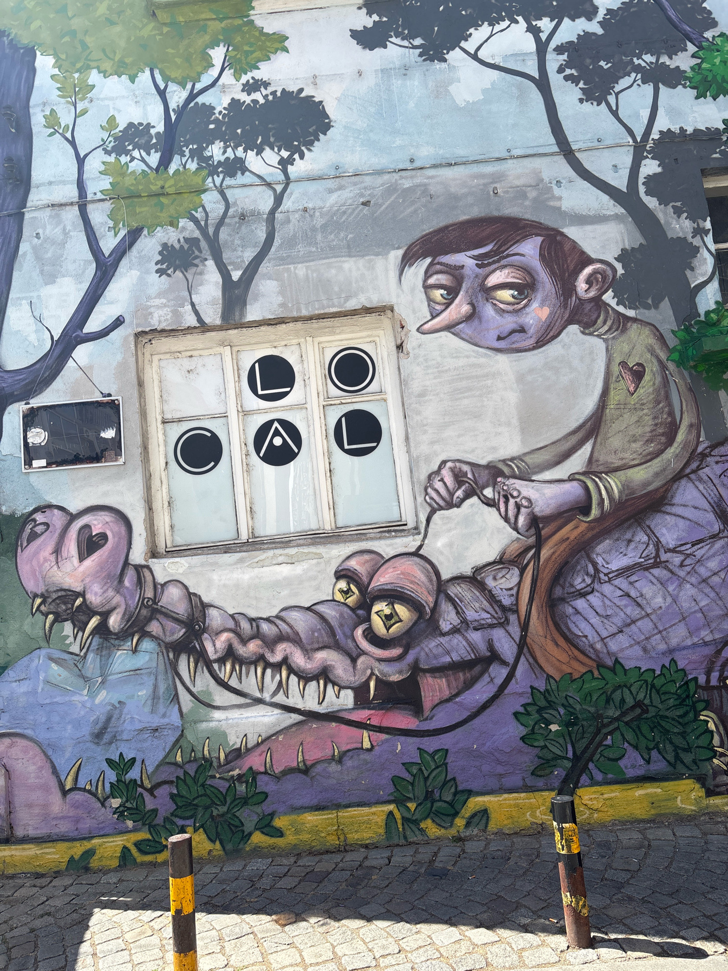 The image depicts a mural painted on the exterior wall of a building in Plovdiv, Bulgaria. The mural features a surreal scene with a purple-skinned character with a long nose riding a stylized purple dragon-like creature. The character is holding reins connected to the creature's mouth. The background of the mural includes stylized trees and a window pane with black circles and triangles, possibly forming letters or symbols. Green foliage is painted at the bottom of the mural, transitioning into a cobblestone street with two bollards. The building's wall shows signs of age and weathering. The overall style is illustrative and cartoonish, with a somewhat melancholic or peculiar mood.