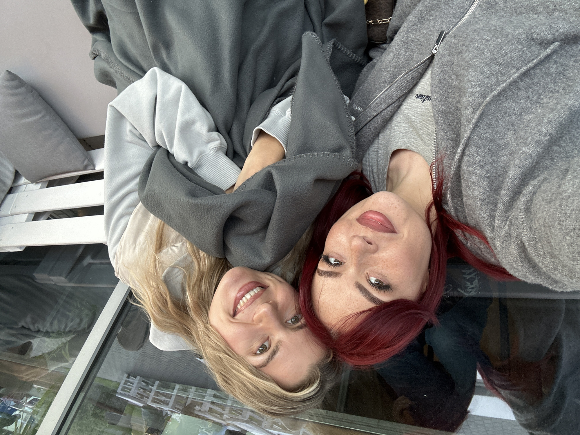 Two women are featured, lying down with their heads close together, smiling brightly at the camera. The woman on the left has blonde hair and is wearing a light grey hoodie, partially covered by a dark grey fleece blanket that also drapes over her arm. The woman on the right has vibrant red hair and is wearing a light grey top under a darker grey zip-up jacket. Both women have warm, engaging smiles, conveying happiness and closeness.
They appear to be lying on a flat, reflective surface, likely glass, which shows blurred reflections of the surrounding environment, possibly outdoor scenery or structures. To the upper left of the blonde woman, a grey pillow rests on a white, slatted piece of furniture, resembling a bench or outdoor sofa. The overall setting suggests a relaxed and cozy atmosphere, possibly on a covered balcony or sunroom in Tolyatti, Russia.
Notable details include the textures of their clothing, from the soft fleece blanket to the ribbed cuffs of the blonde woman's hoodie. Partial text is visible on the red-haired woman's clothing: "ation" on her grey top, and "NSTN" on the zipper pull of her jacket. The lighting indicates it is daytime.