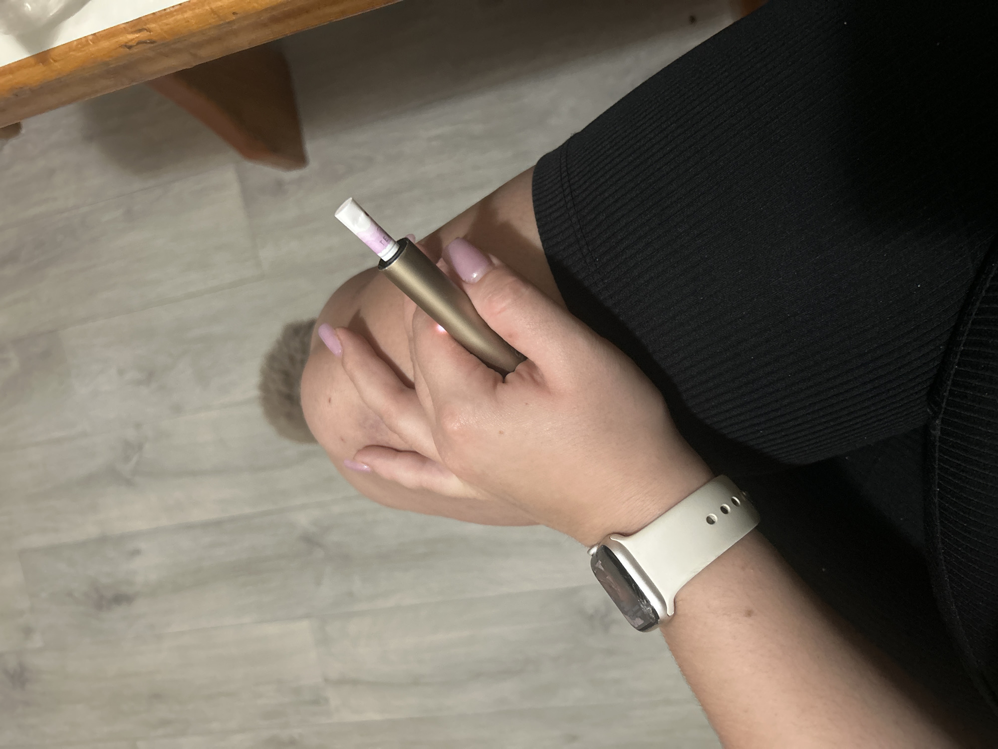 In an indoor setting with a light grey wood-patterned floor, a person's right arm and leg are visible. The individual is wearing a white smartwatch on their left wrist and black ribbed leggings or pants. Their right hand is holding a golden colored electronic smoking device, with a white and pinkish-purple smoking cartridge inserted. The person's fingernails are painted a light pink color. A portion of a wooden table or chair is visible in the upper right corner of the image. There is no information in the image to suggest the location is in the city of Basarabeasca, Moldova.