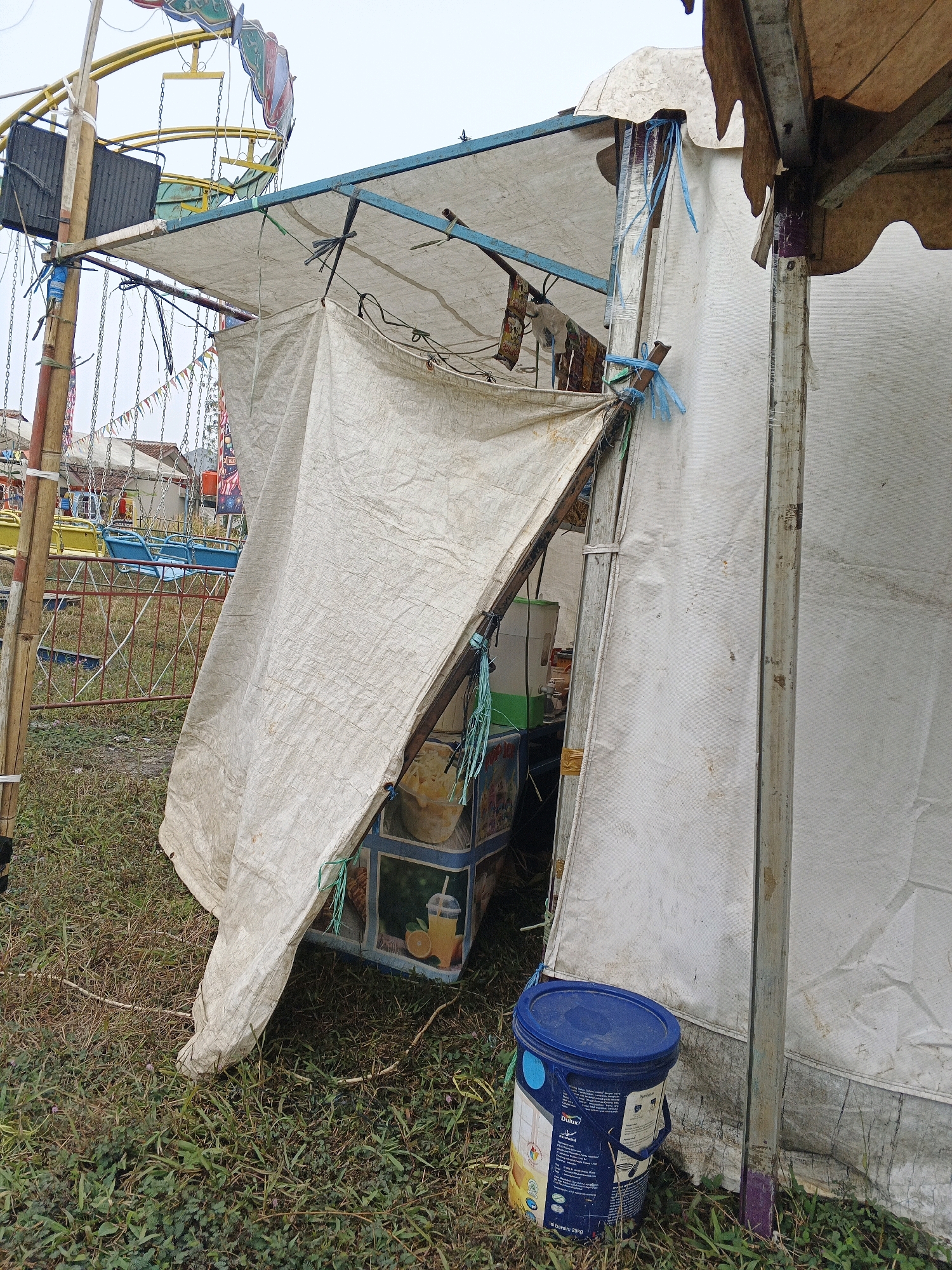 An outdoor scene at a temporary fairground in Sukabumi, Indonesia, features a makeshift food stall constructed with a metal frame and white tarp covering. The stall is positioned on grassy ground, with a portion of a carnival ride, likely a swing ride, visible in the background. The stall displays a refrigerated beverage dispenser and a portable cooler adorned with fruit graphics, suggesting it sells drinks like orange juice. A blue plastic bucket with a yellow lid and branding that reads "Dulux" is placed in front of the stall. The overall impression is one of a humble, temporary setup for a local event.