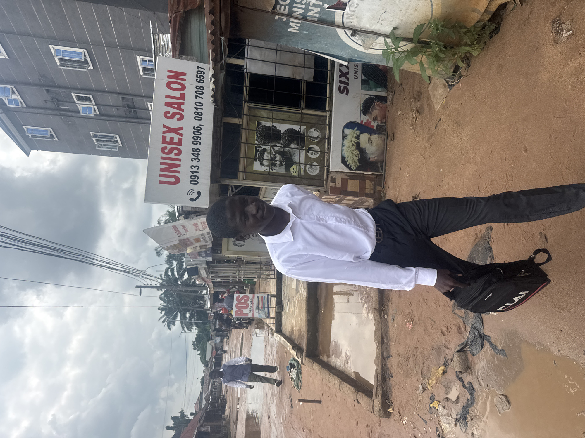 The image depicts a street scene in Ihiagwa, Nigeria, under a cloudy sky. In the foreground, a young man dressed in a white collared shirt, black pants, and a belt is walking towards the camera, holding a black backpack in his right hand. He is on a dirt path that appears to be a road, with several puddles of water and debris scattered around. To the left of the path, there are some structures that look like shops or stalls.
Further down the street, another man in a blue and white shirt and green pants is walking away from the camera. In the background, there are buildings, including a multi-story grey building with blue-trimmed windows on the right, and smaller, more rustic structures on the left. A sign on the grey building reads "UNISEX SALON" in red letters, with phone numbers below. Other signs with images of hairstyles are visible on the storefronts. The overall environment suggests a somewhat unpaved and possibly underdeveloped urban area.