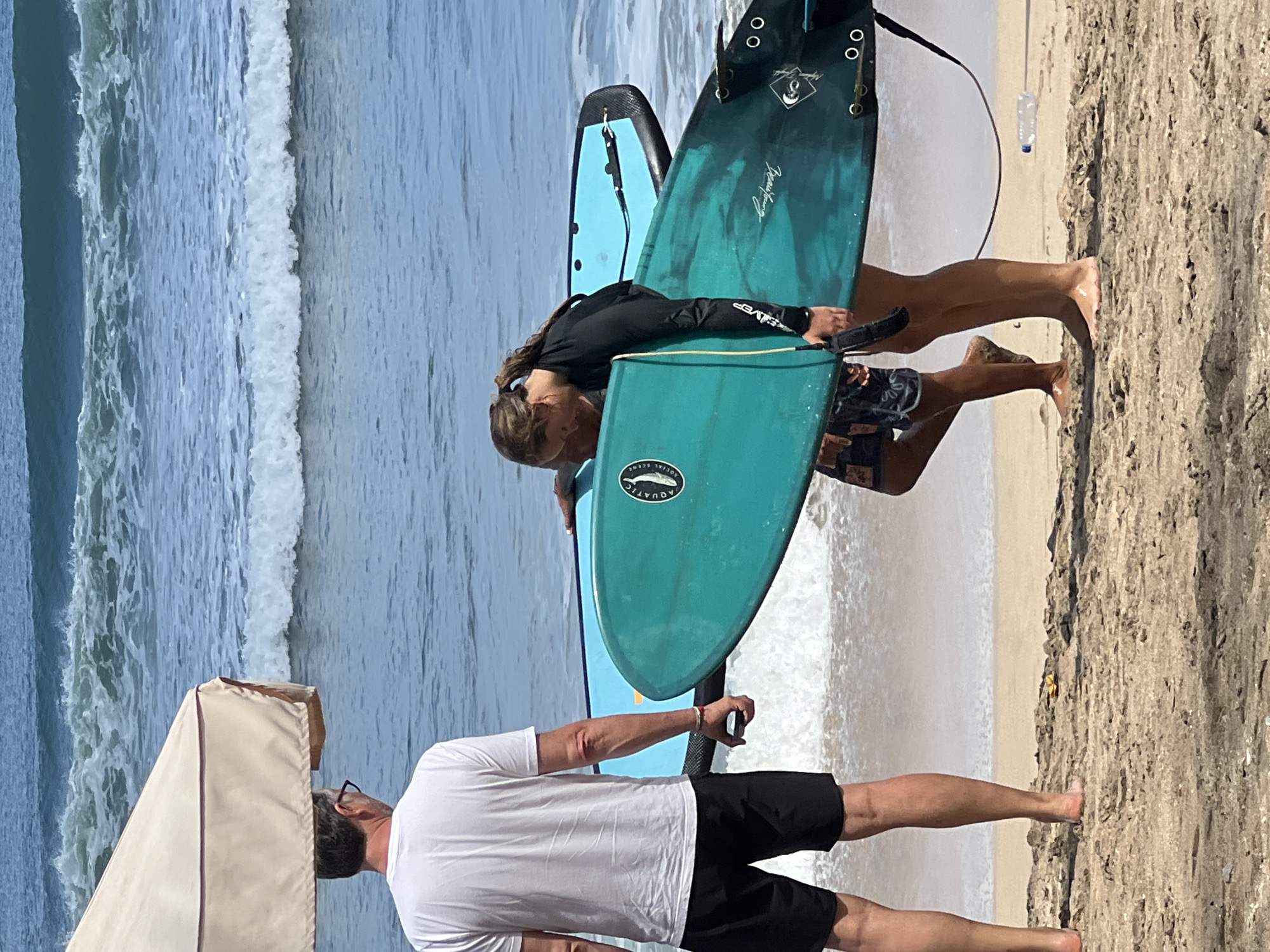 The image depicts a beach scene on a sunny day in Kuta, Indonesia. In the foreground, a man wearing a white t-shirt and black shorts stands with his back to the camera, looking out at the ocean. Beside him, a young woman in a black long-sleeved rash guard and patterned board shorts carries a teal surfboard. Another surfboard, light blue in color, is visible behind the teal one. The ocean in the background has moderate waves breaking on the shore. The beach is sandy, with some debris scattered on the ground, including a plastic water bottle. The overall atmosphere suggests a leisurely day at the beach with surfers preparing to or having finished their session.
