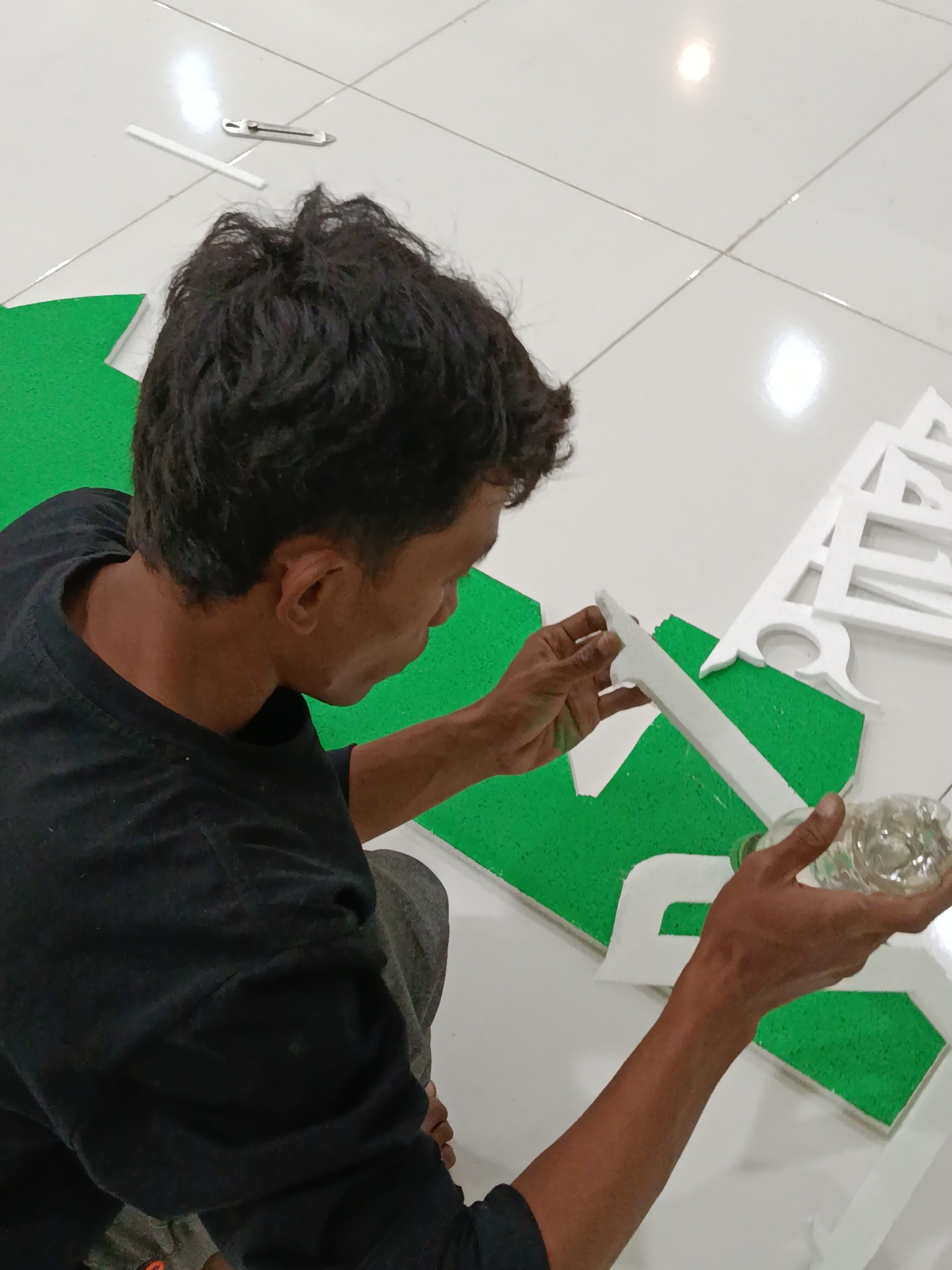 A person is crafting decorations on a white tiled floor in Ramban Wetan, Indonesia. The person, wearing a black t-shirt and gray pants, is crouched down, holding a piece of white foam lettering in their left hand and a clear plastic bottle of liquid in their right hand. The person is in the process of applying the liquid to the foam. Green foam pieces are laid out on the floor, along with other white foam letters. A utility knife and a white stick are also visible on the floor. The lighting suggests an indoor setting.